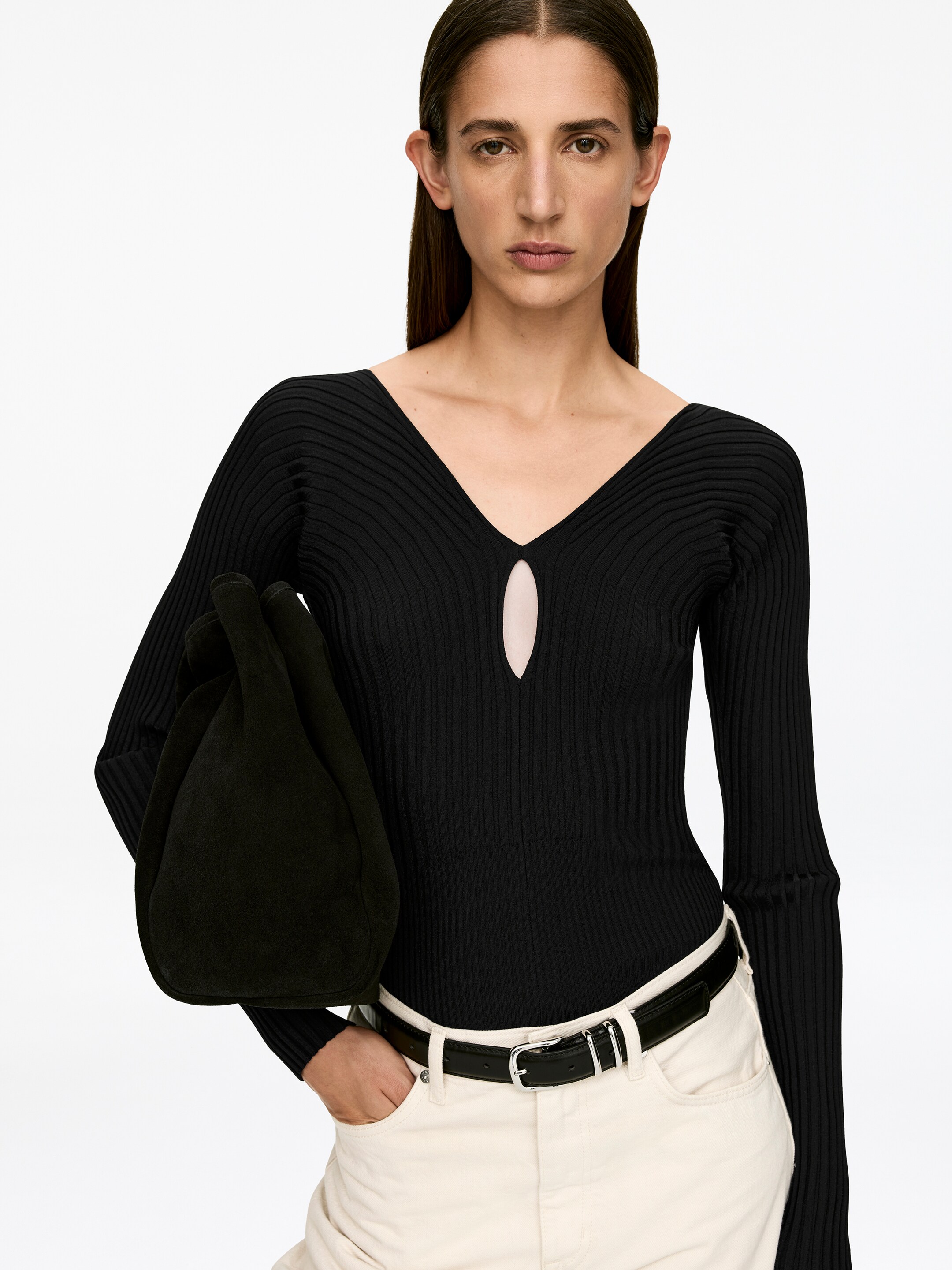 Grotere afbeelding bekijken: Een zwart ribgebreide top, met een diepe V-hals en een sleufopening aan de voorkant, wordt door het model gedragen. De top is ontworpen voor een slanke pasvorm en wordt gestyled met lichtgekleurde broek en een zwarte riem.