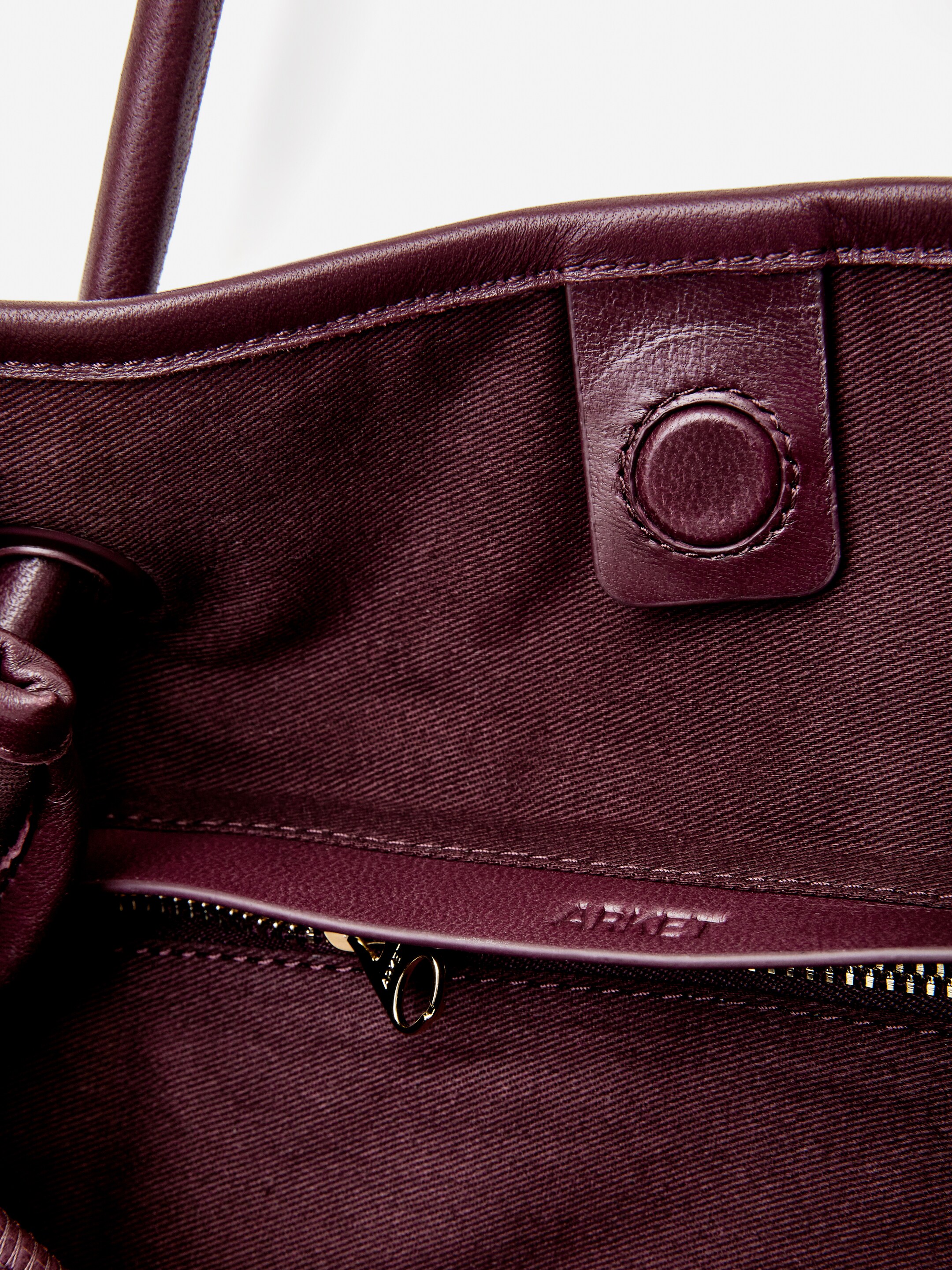Agrandir l'image: Un sac fourre-tout en cuir bordeaux foncé est vu de dessus, montrant son intérieur. Une fermeture à pression magnétique circulaire est visible près de l'ouverture supérieure, et une poche intérieure zippée est également visible, avec une tirette de fermeture éclair de couleur dorée et le nom de la marque "ARKET" embossé sur sa bordure en cuir.