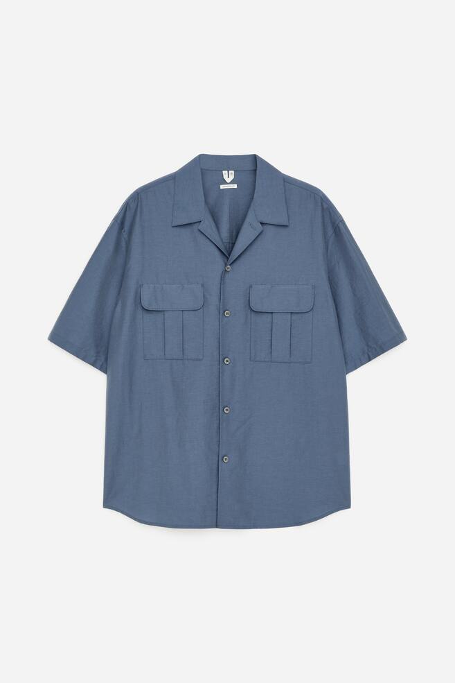 Une chemise bleue à manches courtes est présentée de face, avec un col camp et une patte de boutonnage complète. Deux poches plaquées plissées avec des rabats boutonnés ornent la poitrine. - 1