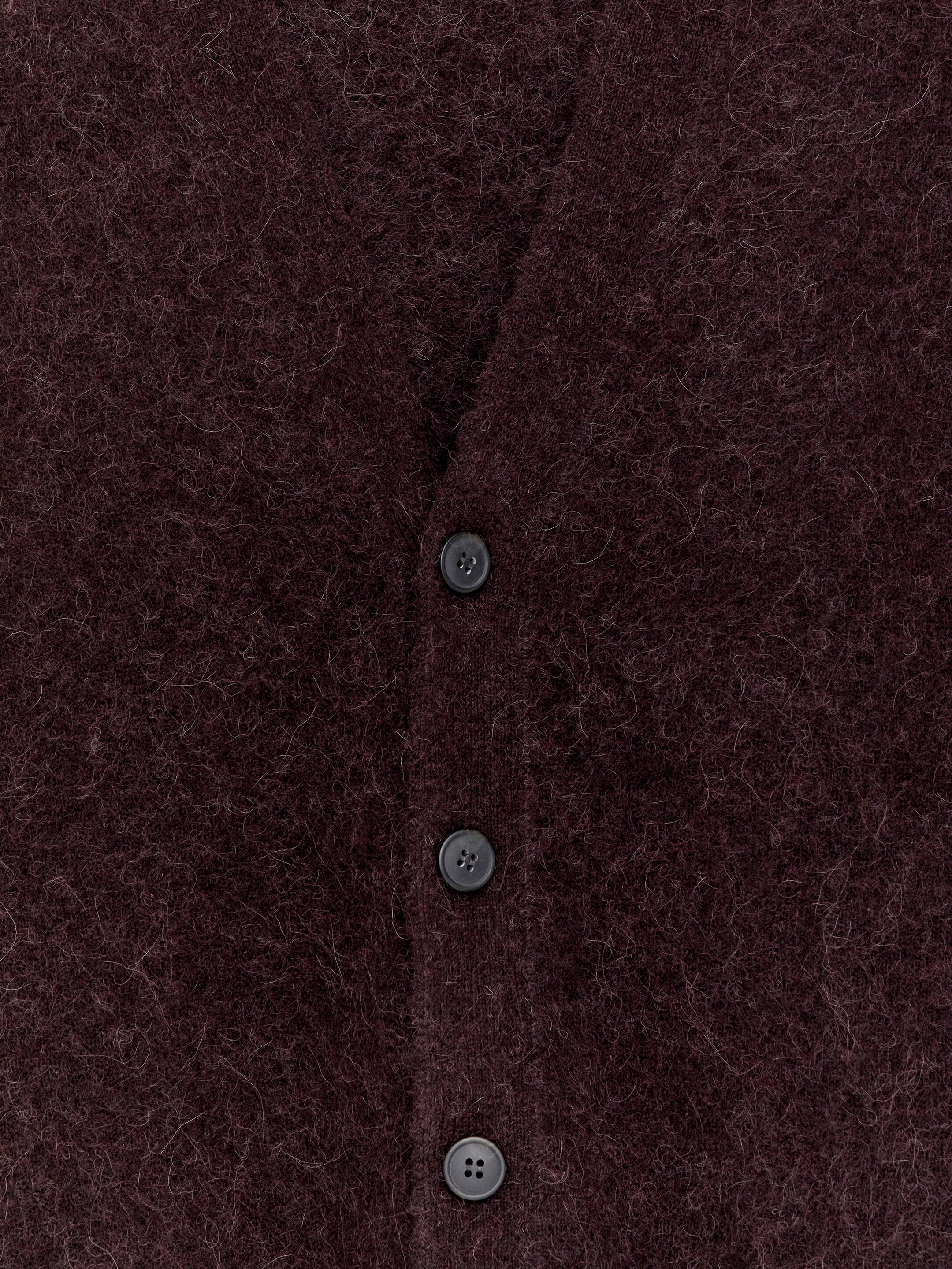 Ver imagem maior: Um casaco de malha vermelho escuro está a ser mostrado de frente, apresentando uma textura peluda proeminente. O seu estilo de decote em V é visível, com três botões escuros a fechar a parte da frente.