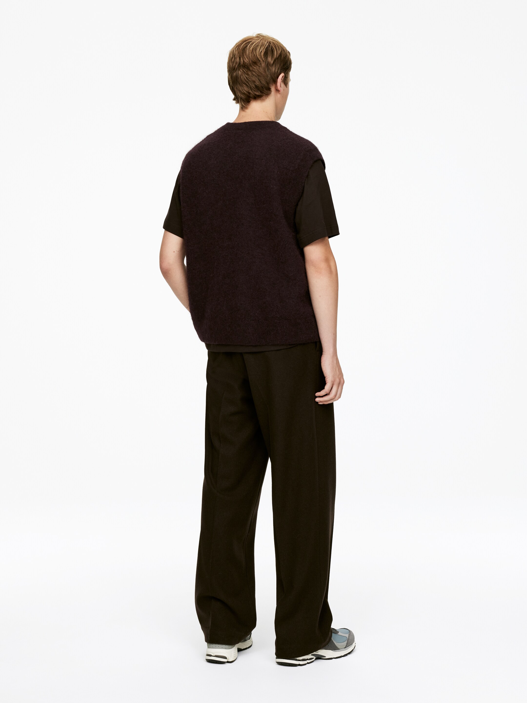 Grotere afbeelding bekijken: Vanuit de achterkant gezien, wordt een donker auberginekleurige, pluizige gebreide sweater-vest gedragen over een donker t-shirt met korte mouwen, gecombineerd met donkerbruine wijde broek.