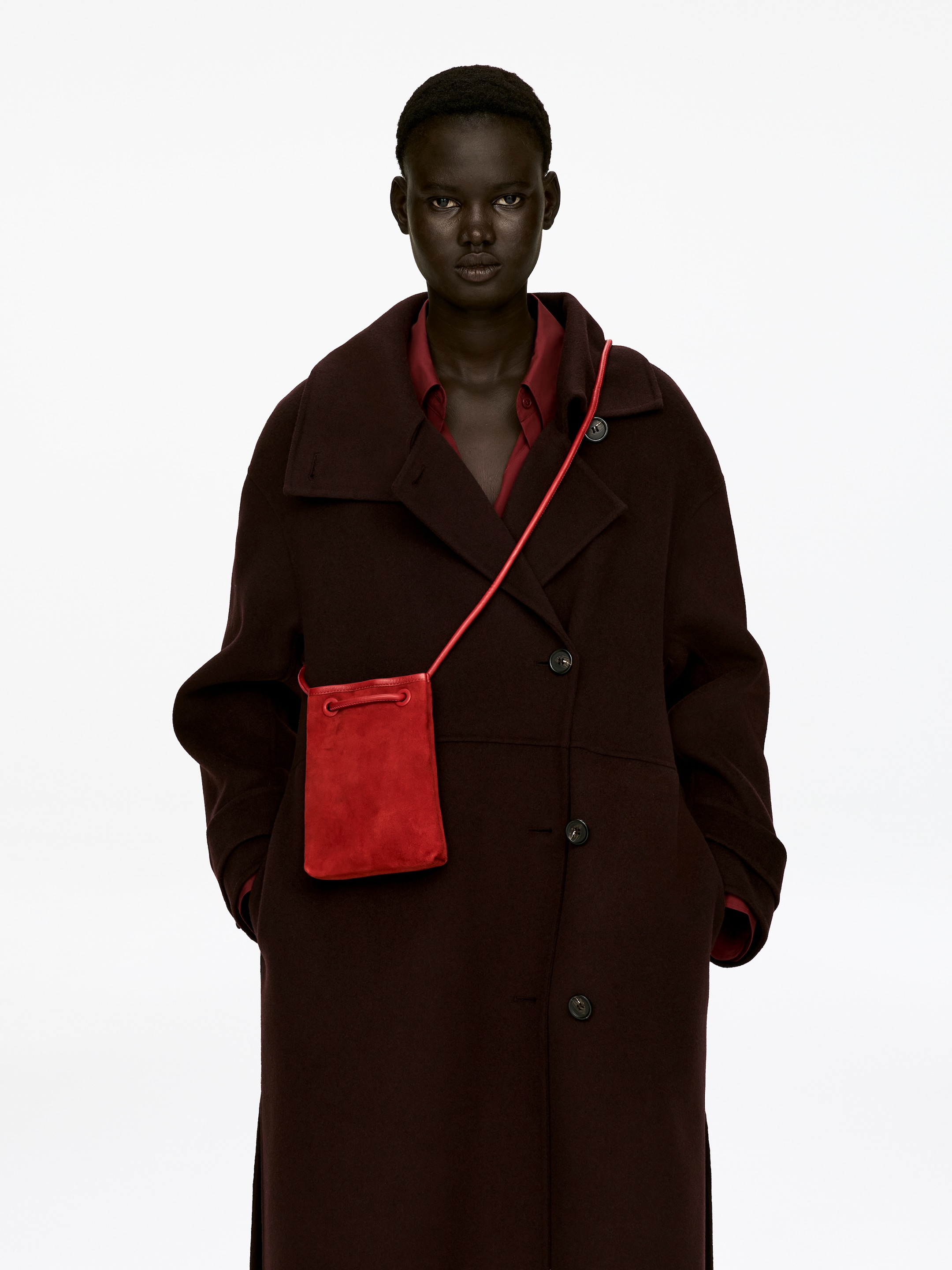 Visualizza immagine più grande: Una borsa a tracolla in pelle scamosciata rossa è indossata dalla modella sopra un cappotto scuro. La sua tracolla tubolare stringe l'apertura della borsa e attraversa la spalla della modella, con la borsa che pende contro il suo fianco.
