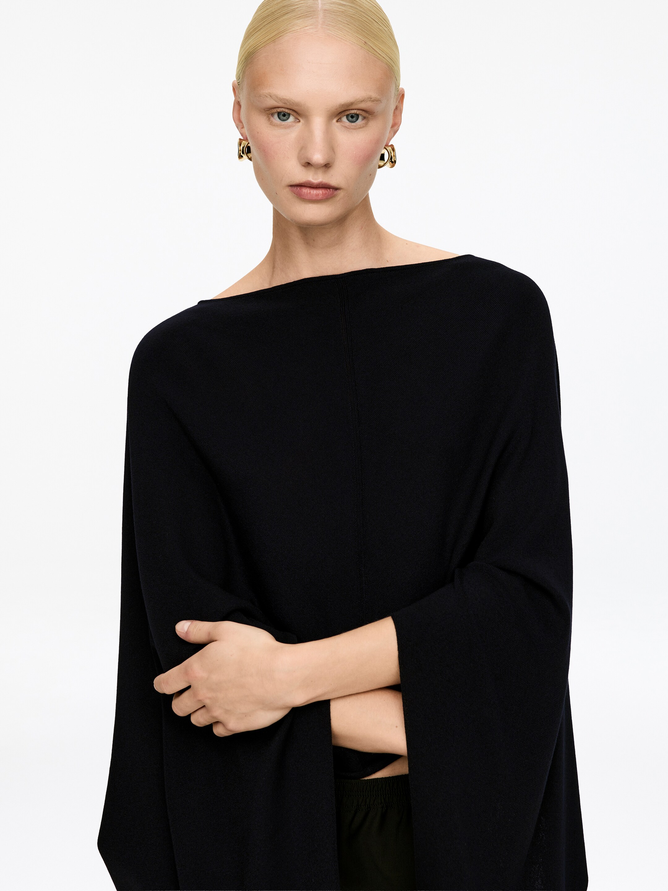 Agrandir l'image: Un haut en tricot noir est tourné vers l'avant, avec une large encolure bateau et des manches larges et décontractées. Une couture verticale descend au centre du devant, et les bras croisés du mannequin mettent en valeur la silhouette drapée du vêtement.