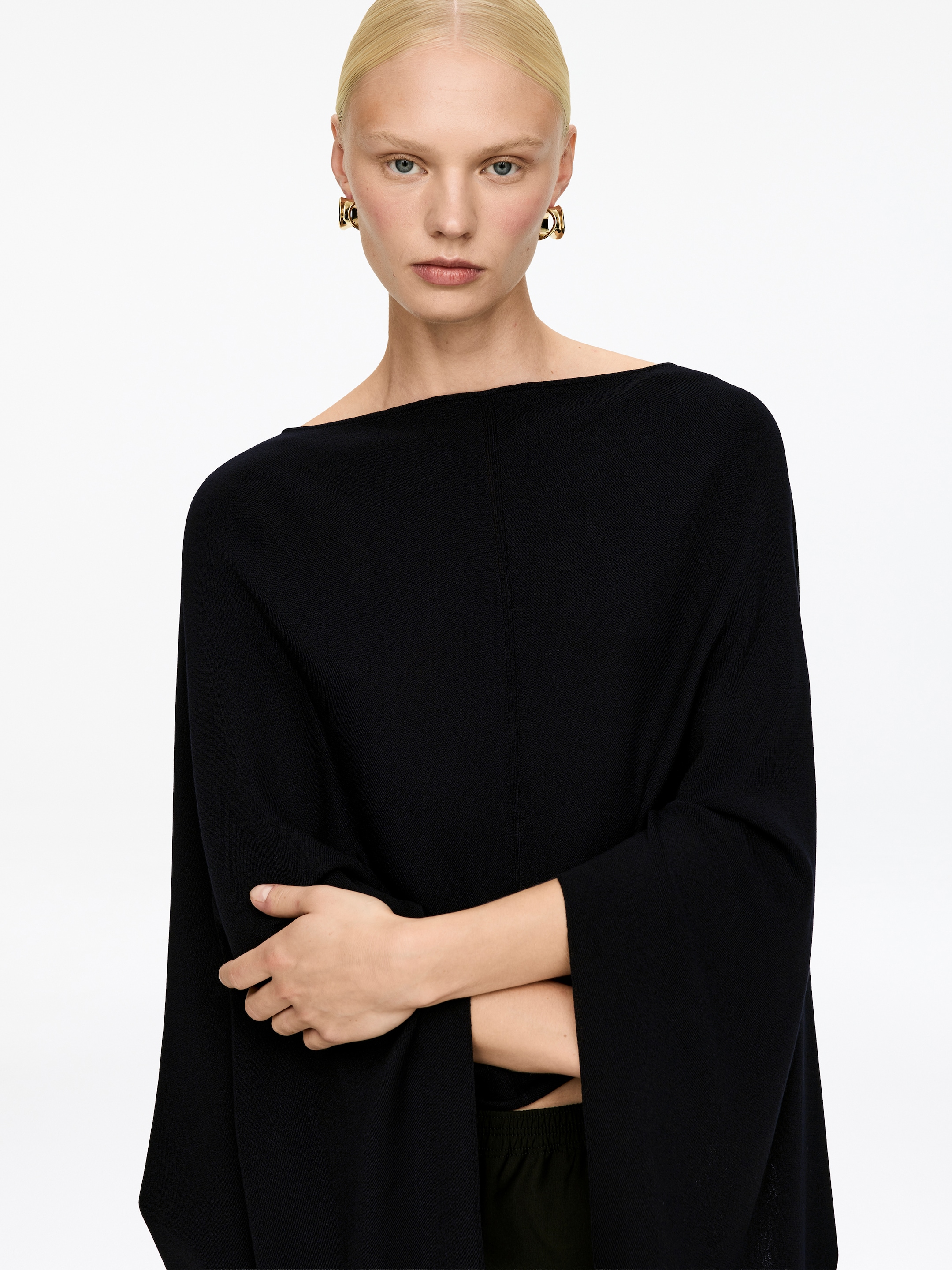 Agrandir l'image: Un haut tricoté noir fait face vers l'avant, avec un large décolleté bateau et des manches larges et décontractées. Une couture verticale descend au milieu du devant, et les bras croisés du mannequin mettent en valeur la silhouette drapée du vêtement.