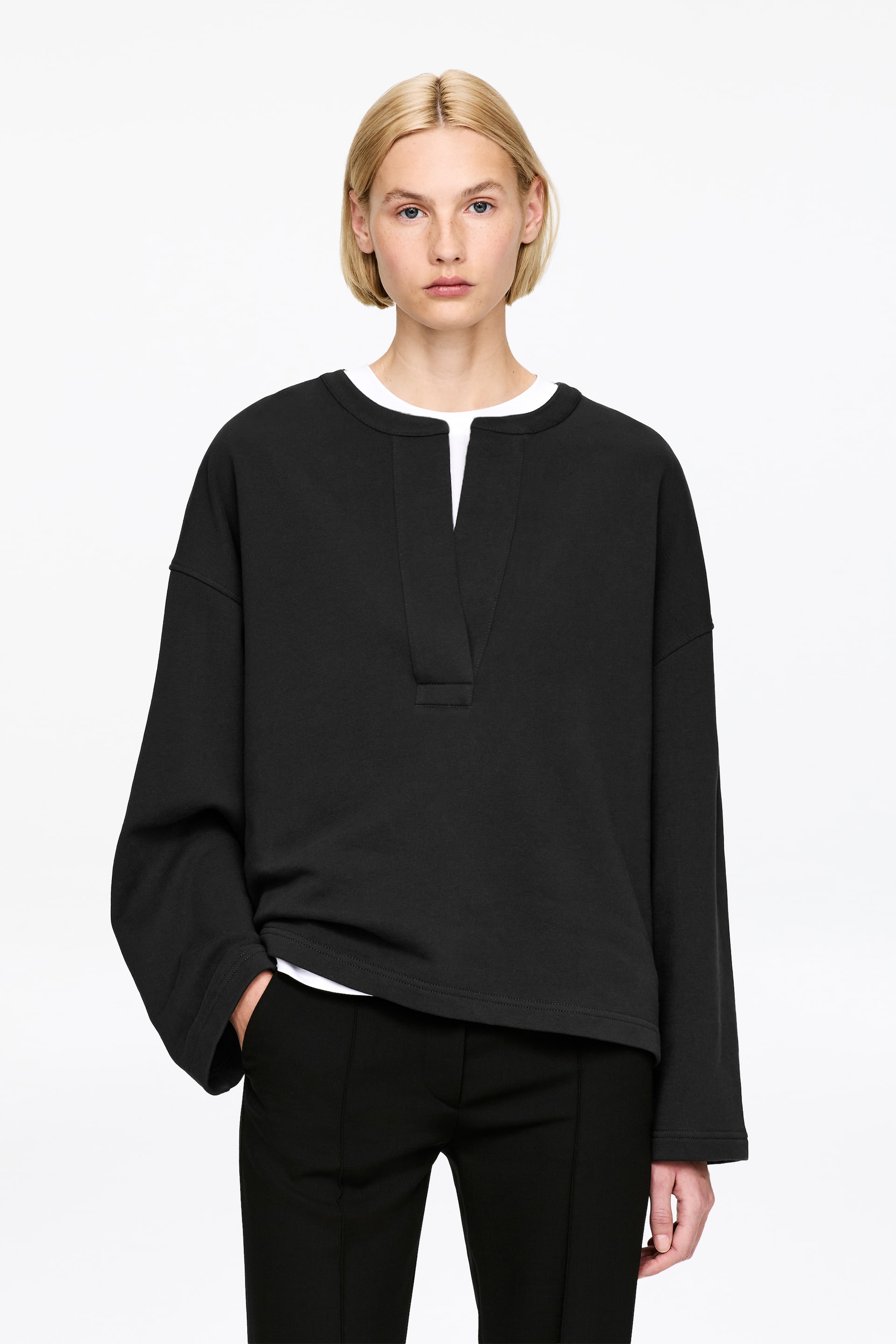 Vezi imaginea mai mare: Un top negru, cu mâneci lungi orientate spre față, prezintă un decolteu rotund cu o deschidere adâncă în formă de V, dezvăluind un tricou alb pe dedesubt. Topul are o croială lejeră, cu mâneci largi, tăiate drept, finisate cu un tiv simplu.