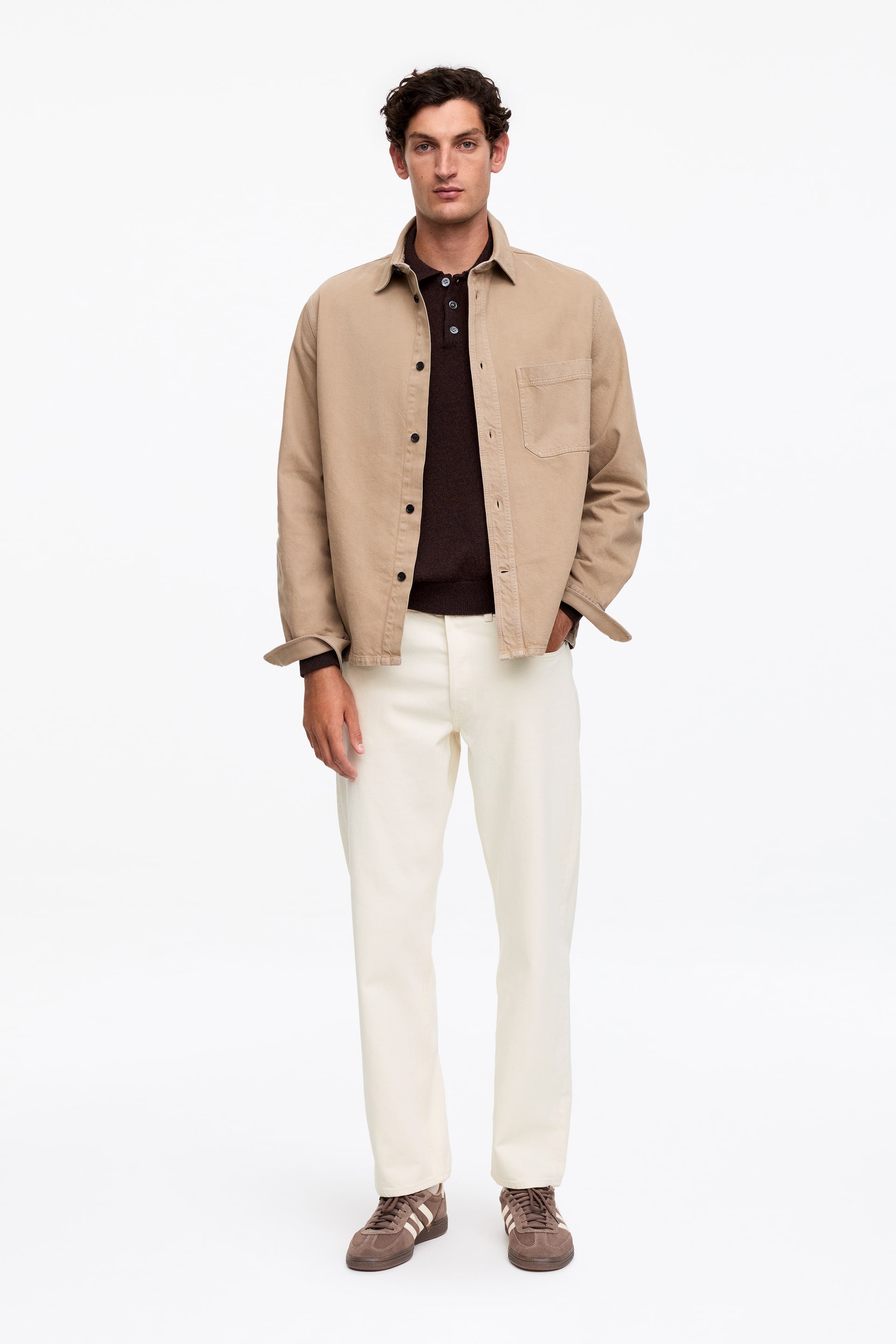 Zobrazit větší obrázek: Outfit obsahuje béžovou košilovou bundu s límečkem, která je otevřeně vrstvená přes tmavě hnědou pletenou polokošili s tříknoflíkovou lištou. Košilová bunda má kapsu na levé straně hrudi a manžetové rukávy, zatímco vzhled doplňují off-white kalhoty s rovnými nohavicemi a hnědé semišové tenisky s bílými bočními pruhy.