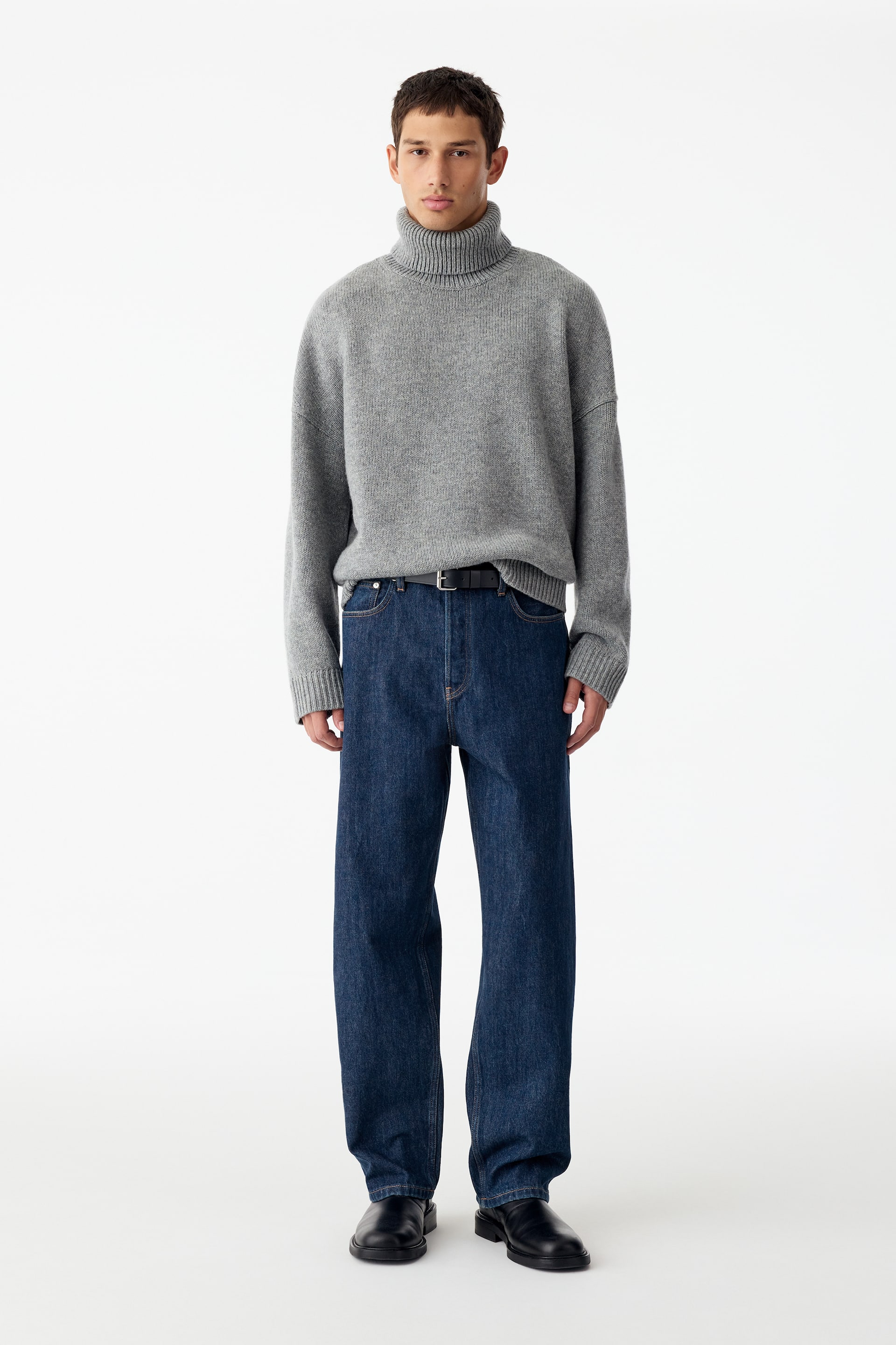 Visa större bild: Mörkblåa vida jeans i denim bärs av modellen, stylade med en grå polotröja och ett svart bälte. De höga jeansen har en lös, hellång passform som faller över svarta skor.