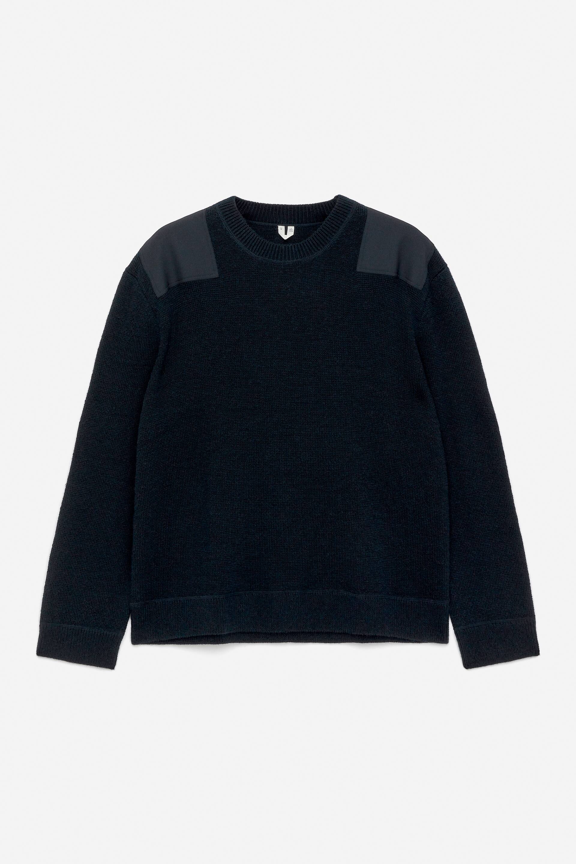 Vis større bilde: En mørk ullgenser med rund hals er vendt mot FORAN, med ribbestrikkede kanter ved halsen, mansjettene og nederkanten, og vevde lapper på skuldrene.