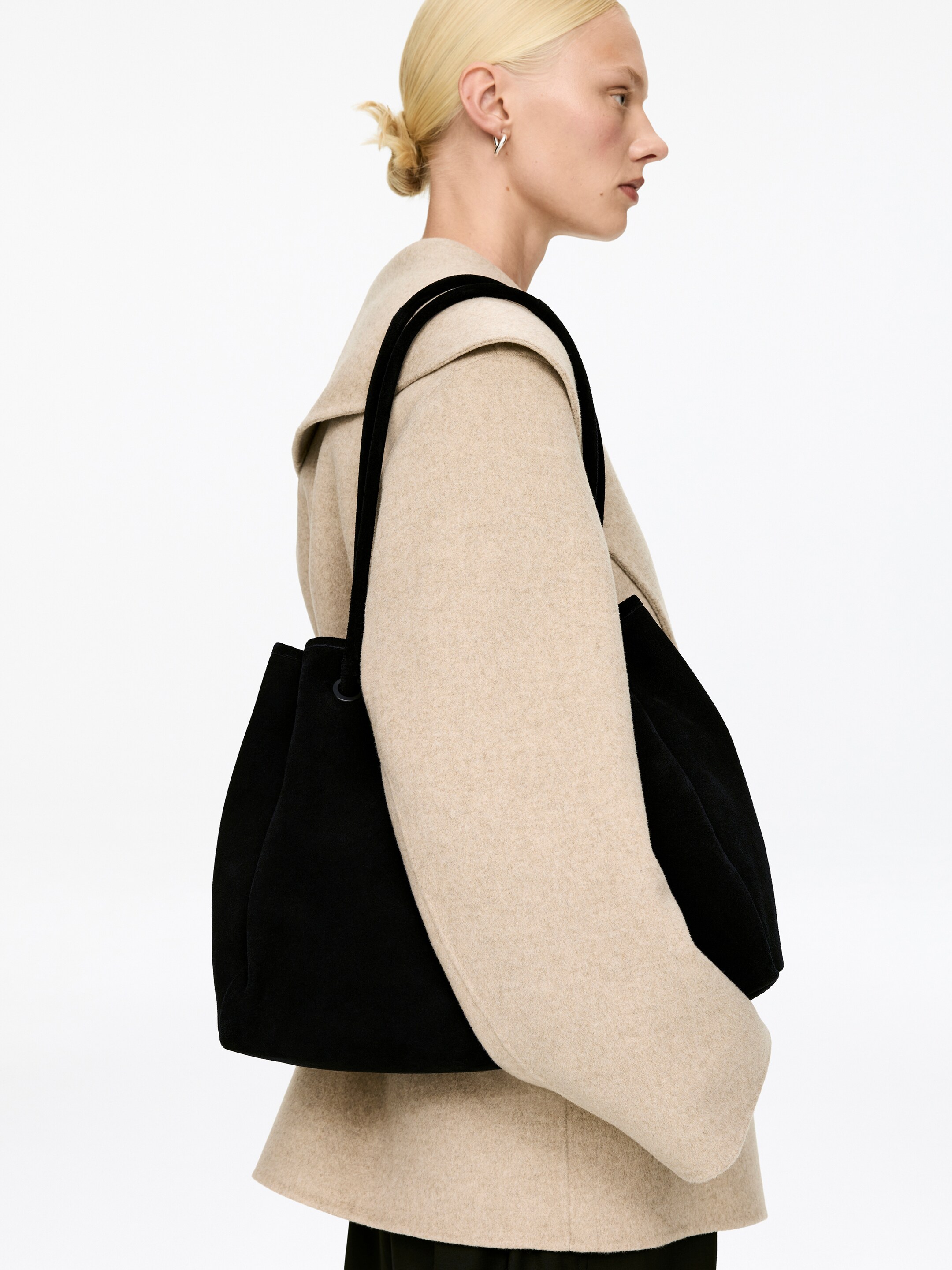 Agrandir l'image: Un sac seau noir et souple est porté sur l'épaule, reposant contre un manteau beige. Il présente des sangles larges et intégrées ainsi qu'une fermeture à cordon visible en haut.