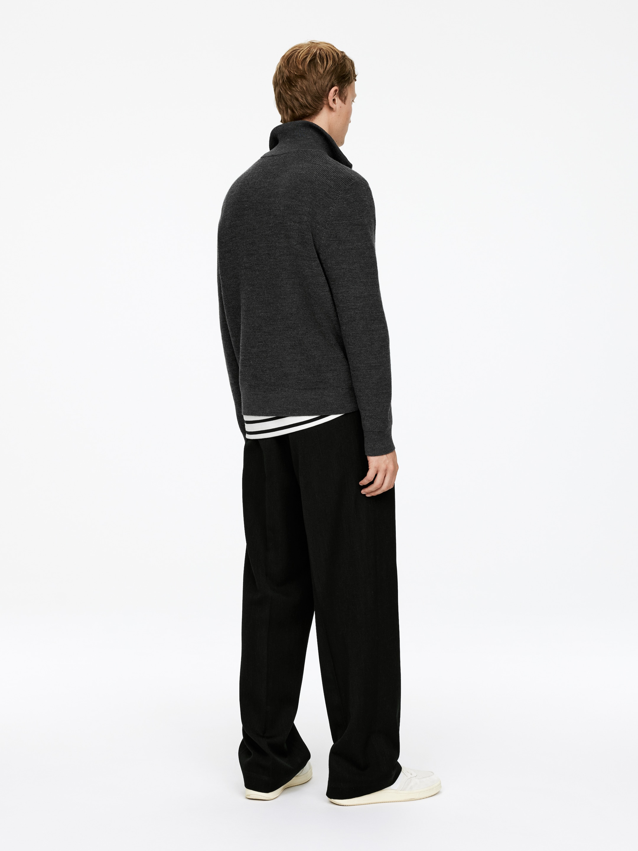 Visualizza immagine più grande: Un maglione a collo alto in maglia grigio scuro è indossato, caratterizzato da un colletto a coste e maniche lunghe, con una camicia a righe bianche e nere che sbuca da sotto. È abbinato a pantaloni neri a gamba larga che cadono su sneakers bianche, il tutto visto da un'angolazione posteriore e laterale.