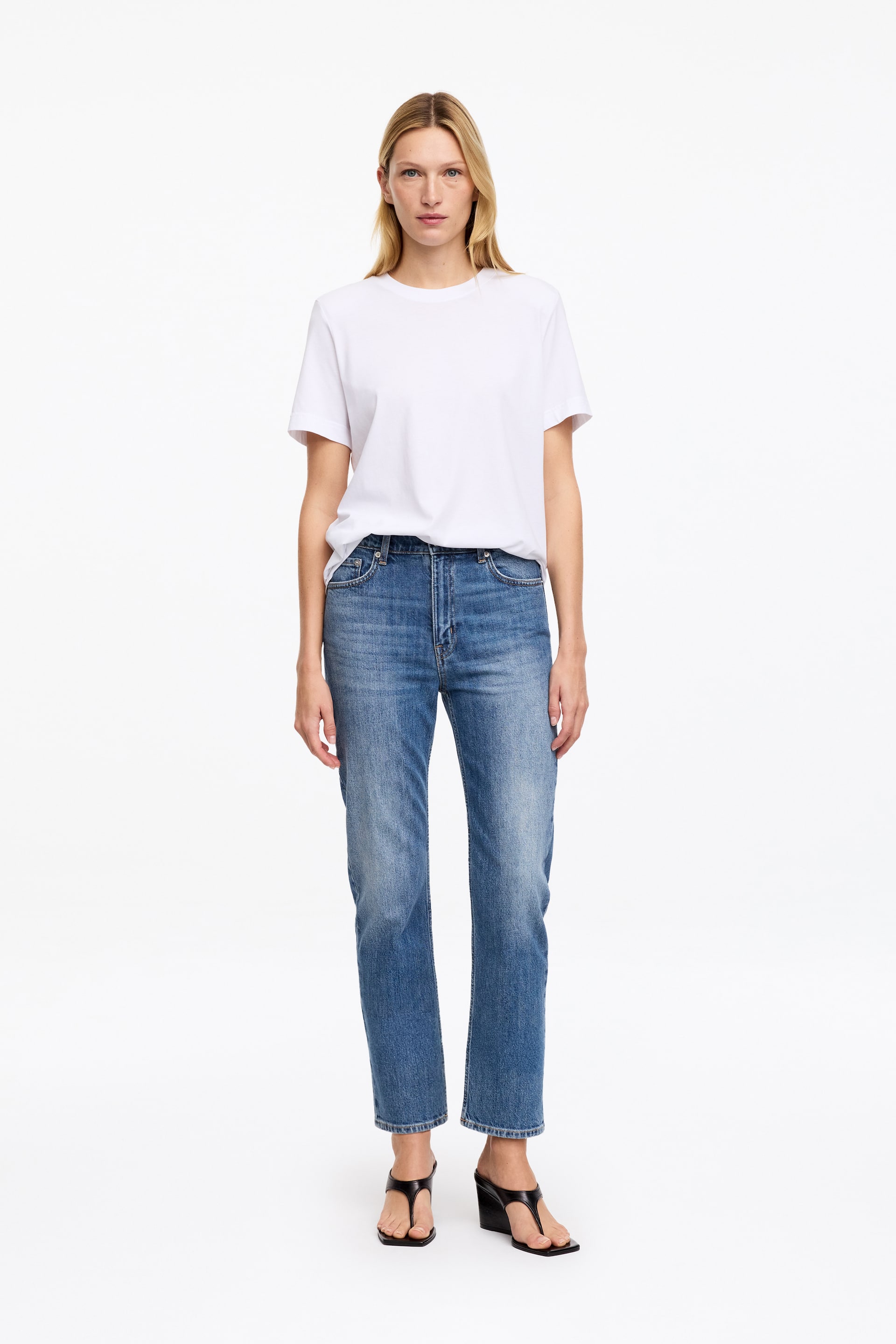 Grösseres Bild ansehen: Blaue Cropped-Jeans aus Denim mit mittlerer Bundhöhe und schlanker Silhouette werden vom Model getragen. Die Jeans sind mit einem teilweise in den Bund gesteckten weissen T-Shirt und schwarzen Keilsandaletten kombiniert.