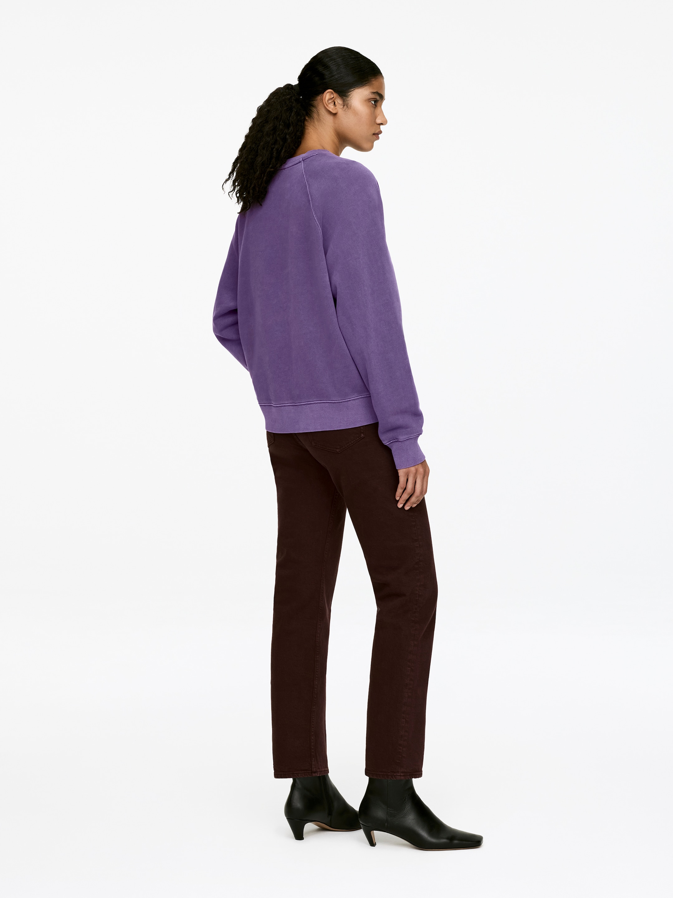 Grösseres Bild ansehen: Ein lilafarbenes Sweatshirt mit Rundhalsausschnitt, Raglanärmeln, gerippten Abschlüssen und normaler Passform wird vom Model getragen. Das Model präsentiert das Sweatshirt, kombiniert mit dunklen Hosen und schwarzen Ankle Boots.