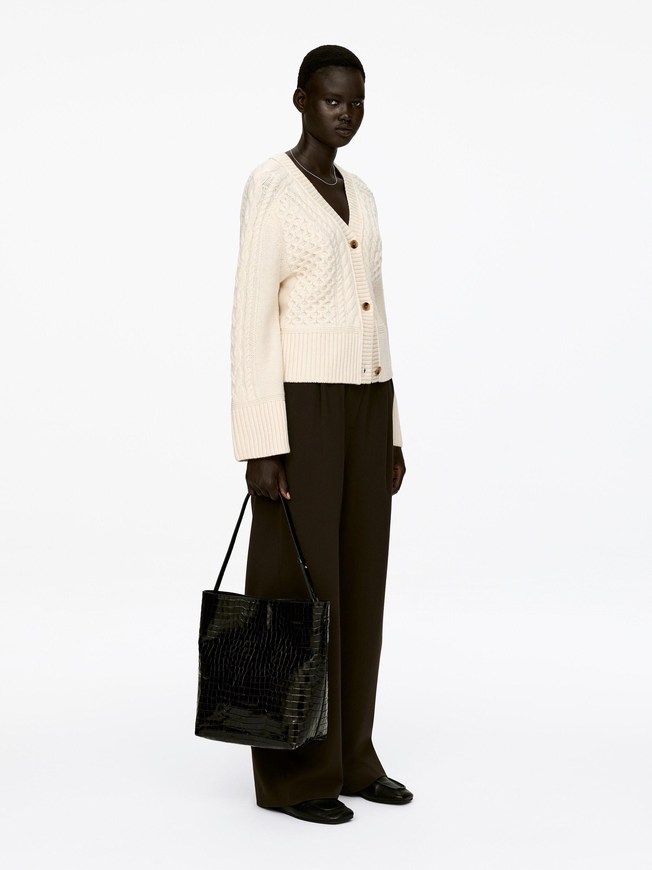 Agrandir l'image: Un cardigan en maille torsadée crème avec un col en V, une fermeture à trois boutons et des bords côtelés est porté par le mannequin. Il est attaché avec deux boutons, porté sur un pantalon foncé, et un collier en argent est visible au niveau du col en V.