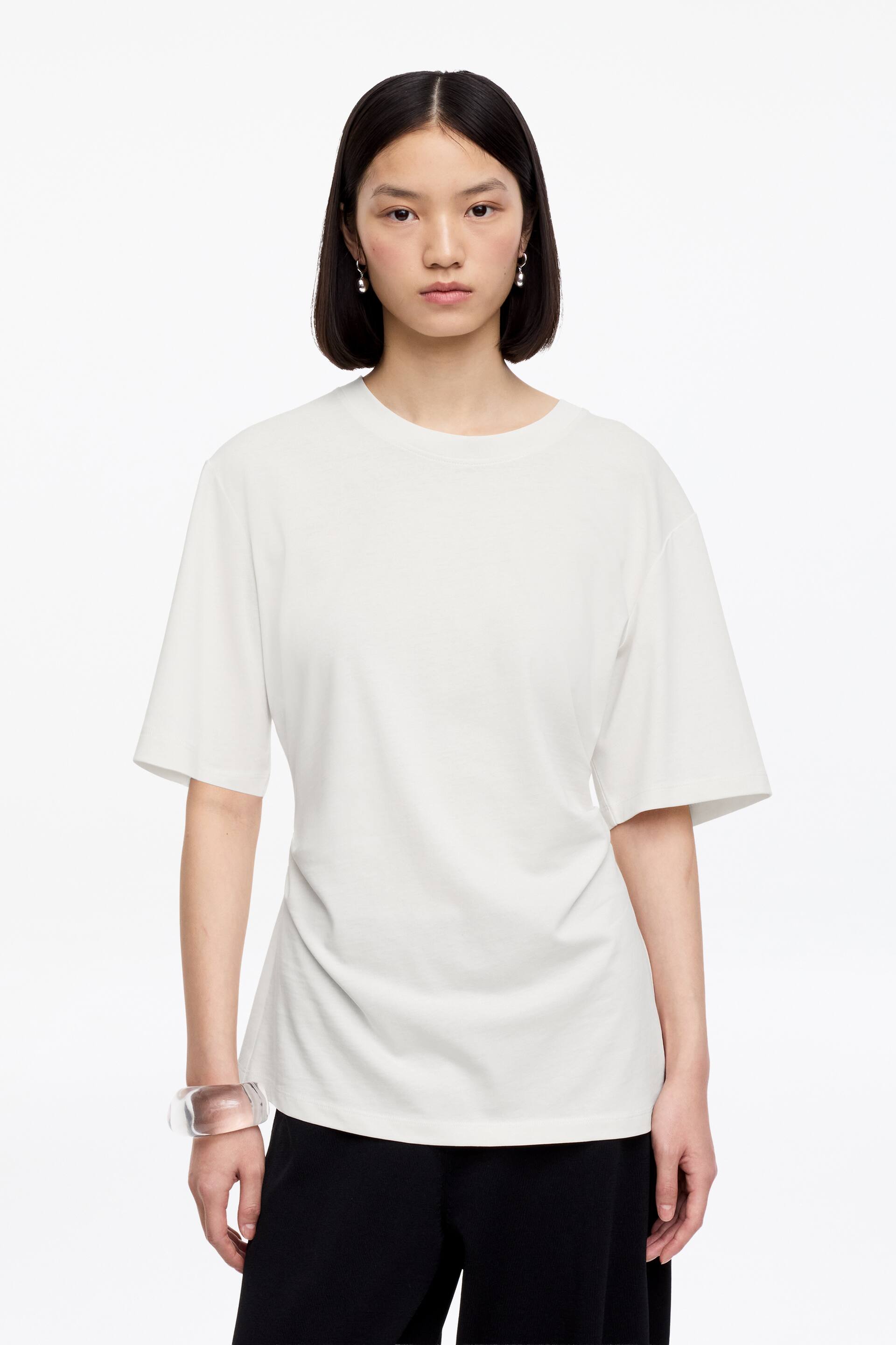 Vezi imaginea mai mare: Un tricou alb prezintă un guler rotund și mâneci scurte. Are un croi lejer din față, cu un material neted care se așază lejer până la tivul drept.