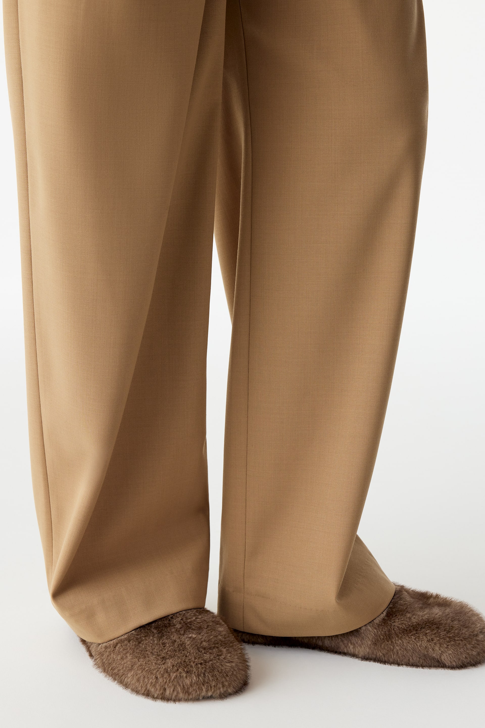View larger image: Brown, fluffy slippers with a rounded shape are being worn by the model. The slippers are styled with tan wide-leg trousers that drape over the ankles.