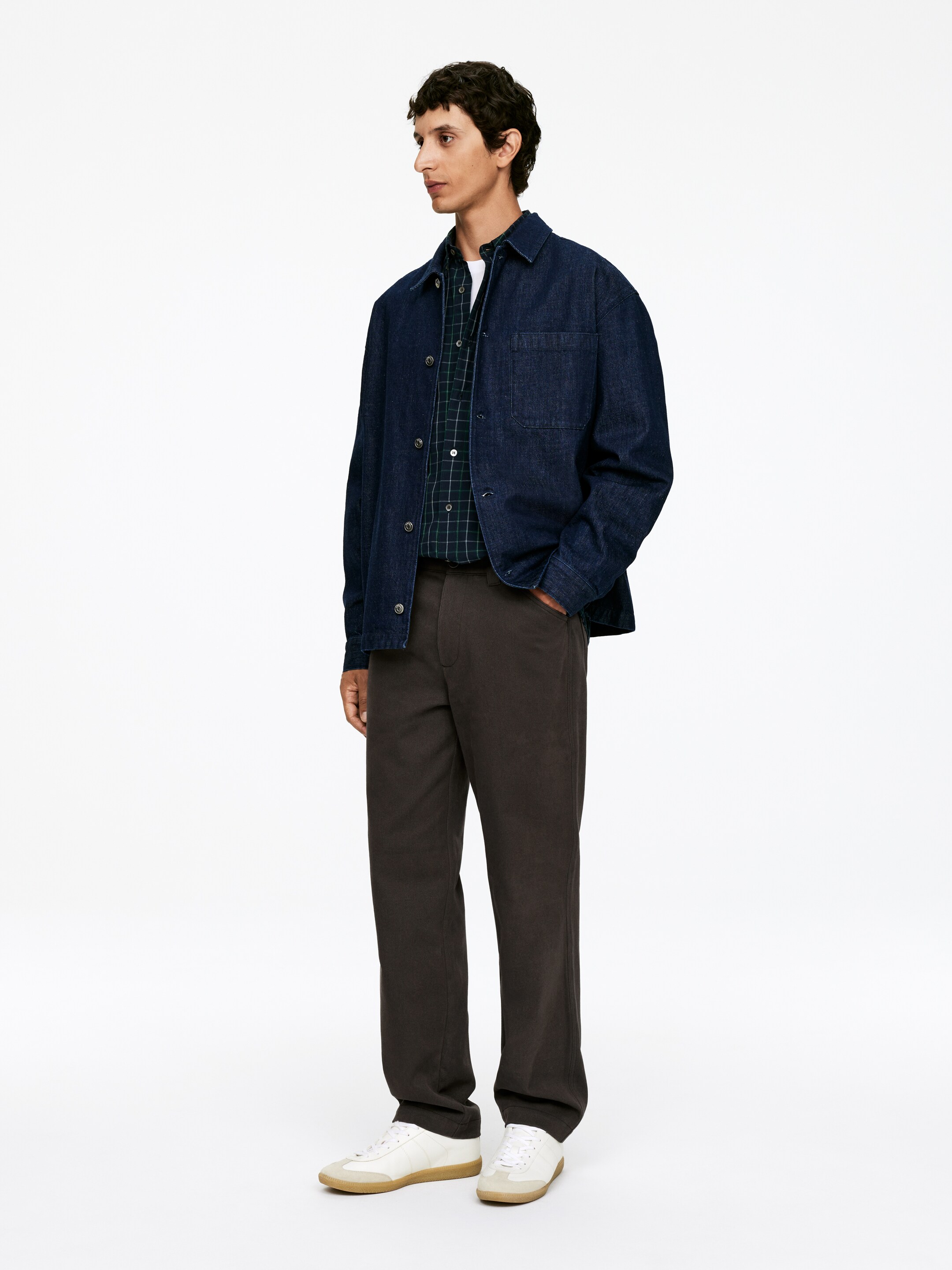 Visualizza immagine più grande: Una camicia di jeans blu scuro viene indossata aperta, rivelando una camicia a quadri verde scuro e blu sotto. La camicia di jeans presenta un colletto classico e una singola tasca a toppa sul petto, ed è abbinata a pantaloni grigio scuro a gamba dritta e sneakers bianche a basso profilo con suole marrone chiaro.