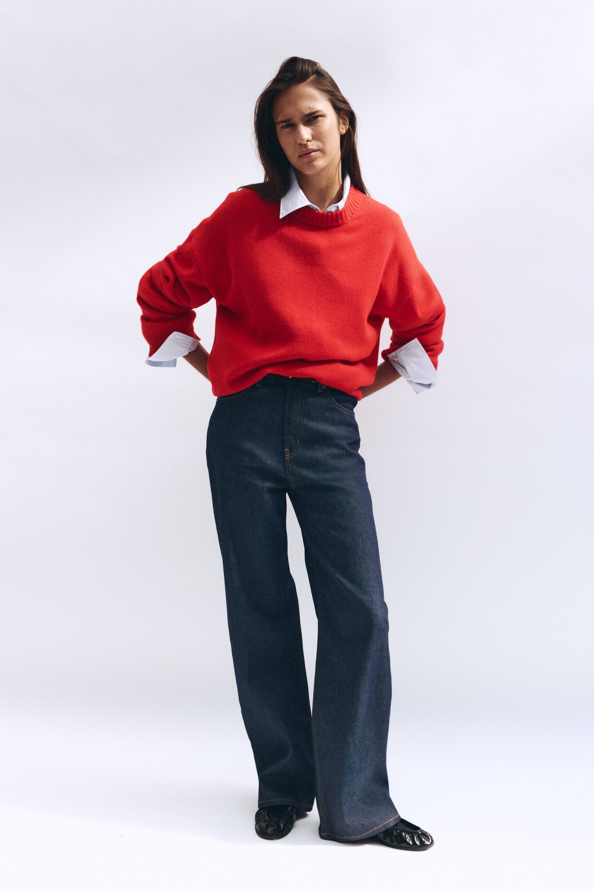 Ver imagem maior: Uma modelo está virada para a frente, a usar uma camisola vermelha de gola redonda, de corte descontraído, com ombros descaídos, sobreposta a uma camisa clássica com colarinho, com os punhos dobrados para trás. A peça superior é combinada com jeans de ganga escura, de cintura subida, com corte slim e perna à boca de sino, finalizados com mocassins em pele envernizada preta.