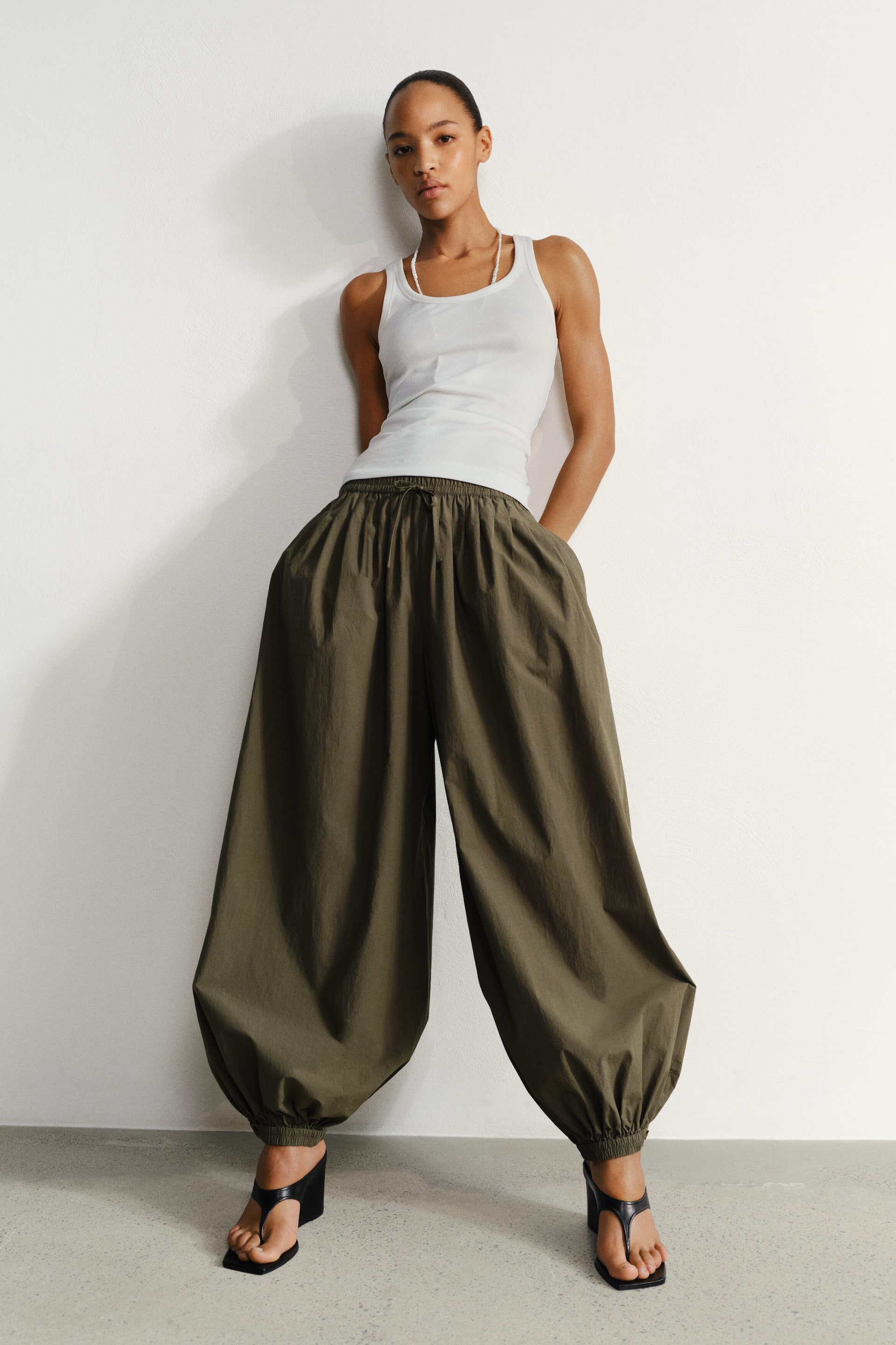 Vezi imaginea mai mare: Pantalonii verde măsliniu prezintă un design voluminos, cu craci largi, având o talie cu șnur vizibil, buzunare laterale și glezne strânse cu elastic, creând o siluetă tip balon.