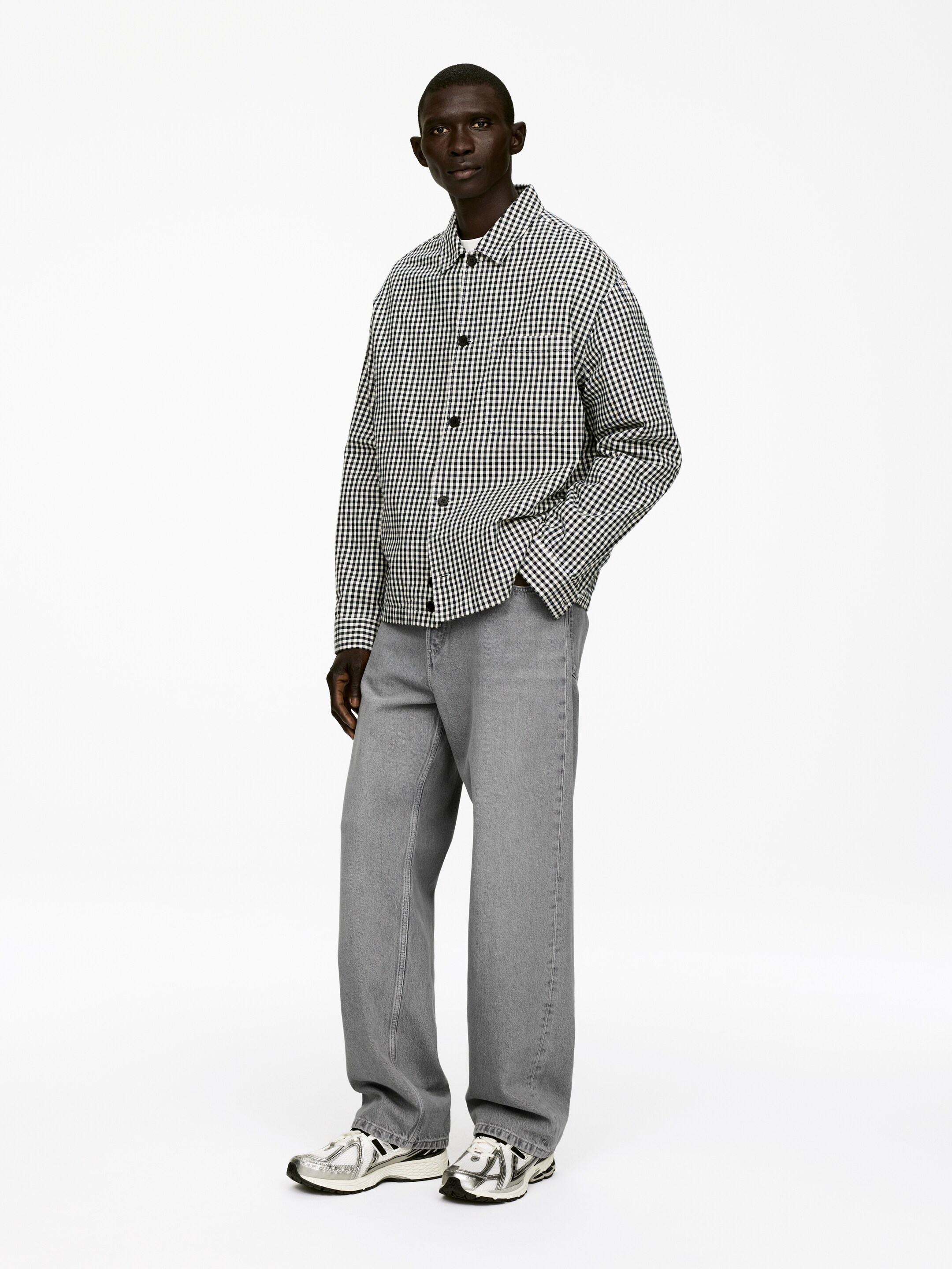 Agrandir l'image: Une surchemise à carreaux noirs et blancs surdimensionnée avec des boutons foncés est portée ouverte sur un t-shirt blanc à col rond. Elle est assortie à un jean gris à jambes larges et amples et des baskets athlétiques de couleur argentée.