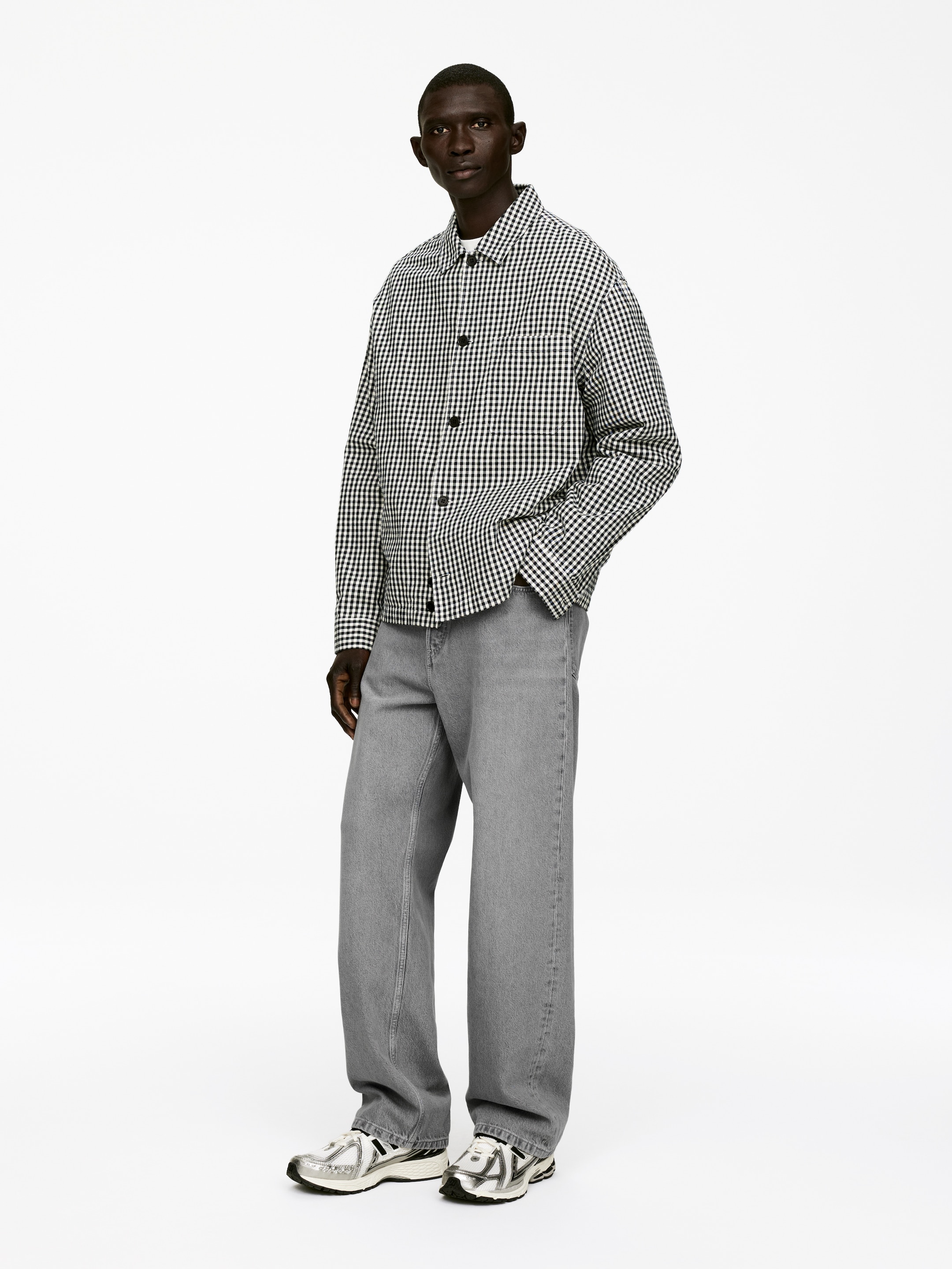 Ingrandisci l'immagine: Una giacca camicia a quadri bianchi e neri oversize con bottoni scuri è indossata aperta sopra una t-shirt bianca con scollo a giro. È abbinata a jeans grigi a gamba larga e sneakers sportive color argento.