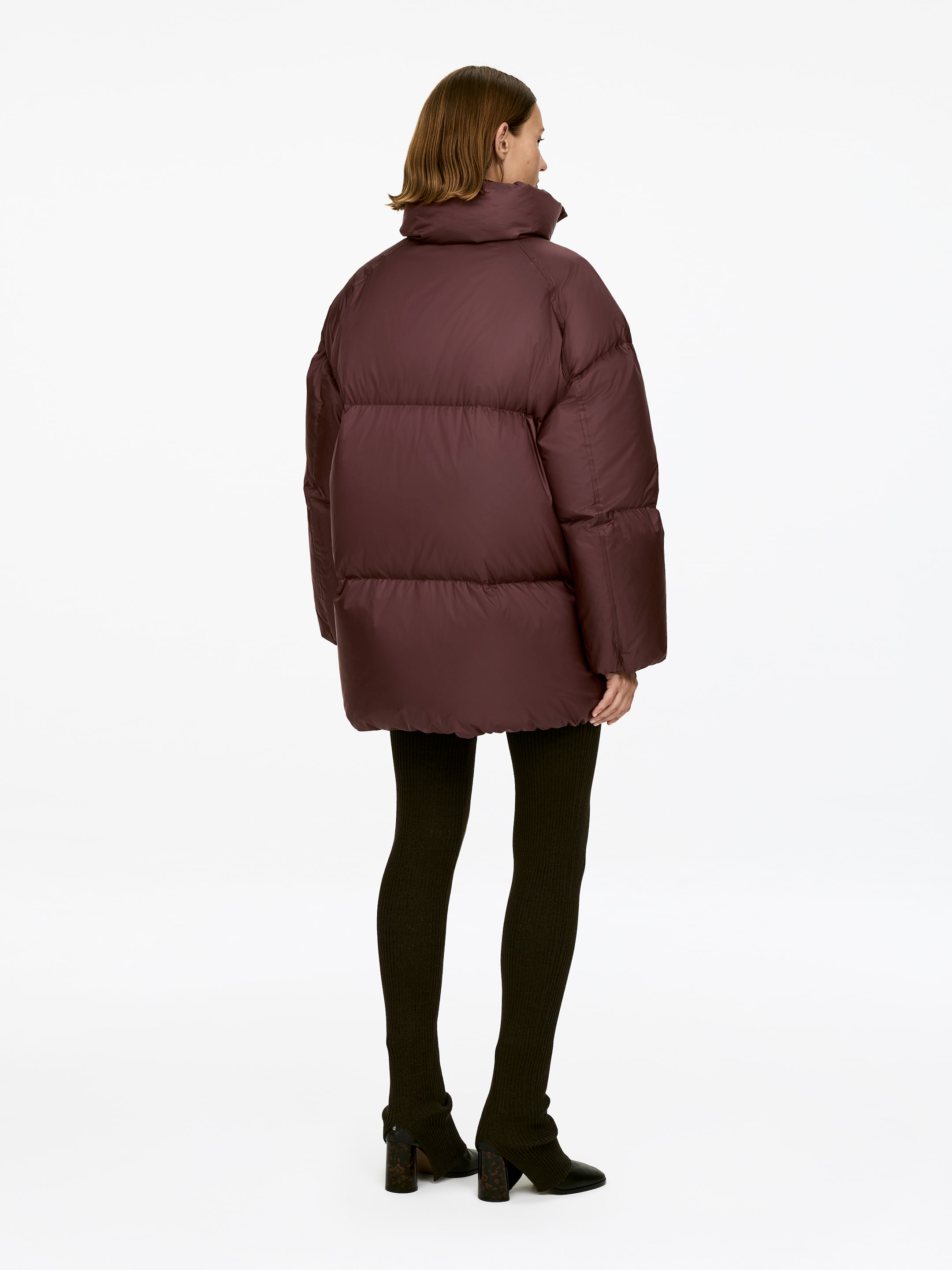 View larger image: Upcycled Down Puffer Jacket. The oversized, dark plum puffer jacket, featuring a high stand collar and quilted design, is being worn by the model from a rear view.