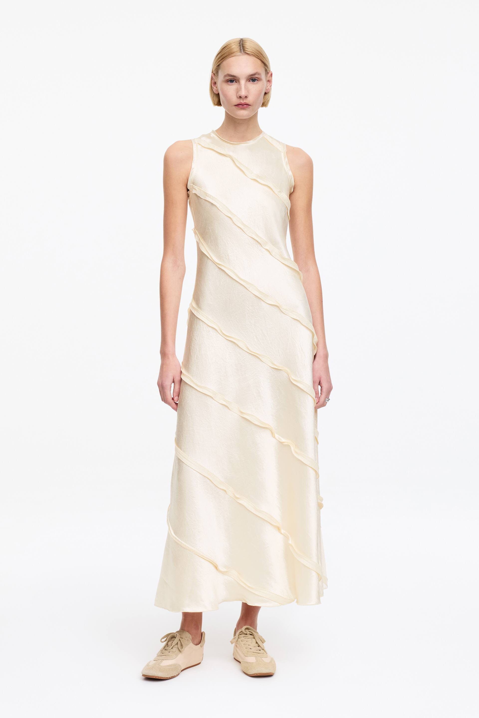 Se større billede: En cremefarvet ærmeløs lang kjole med en rund halsudskæring og et tekstureret, skinnende stof. Diagonale striber af matchende stof er påsyet foran, der strækker sig fra skulderen til sømmen, hvilket skaber et bølget mønster.
