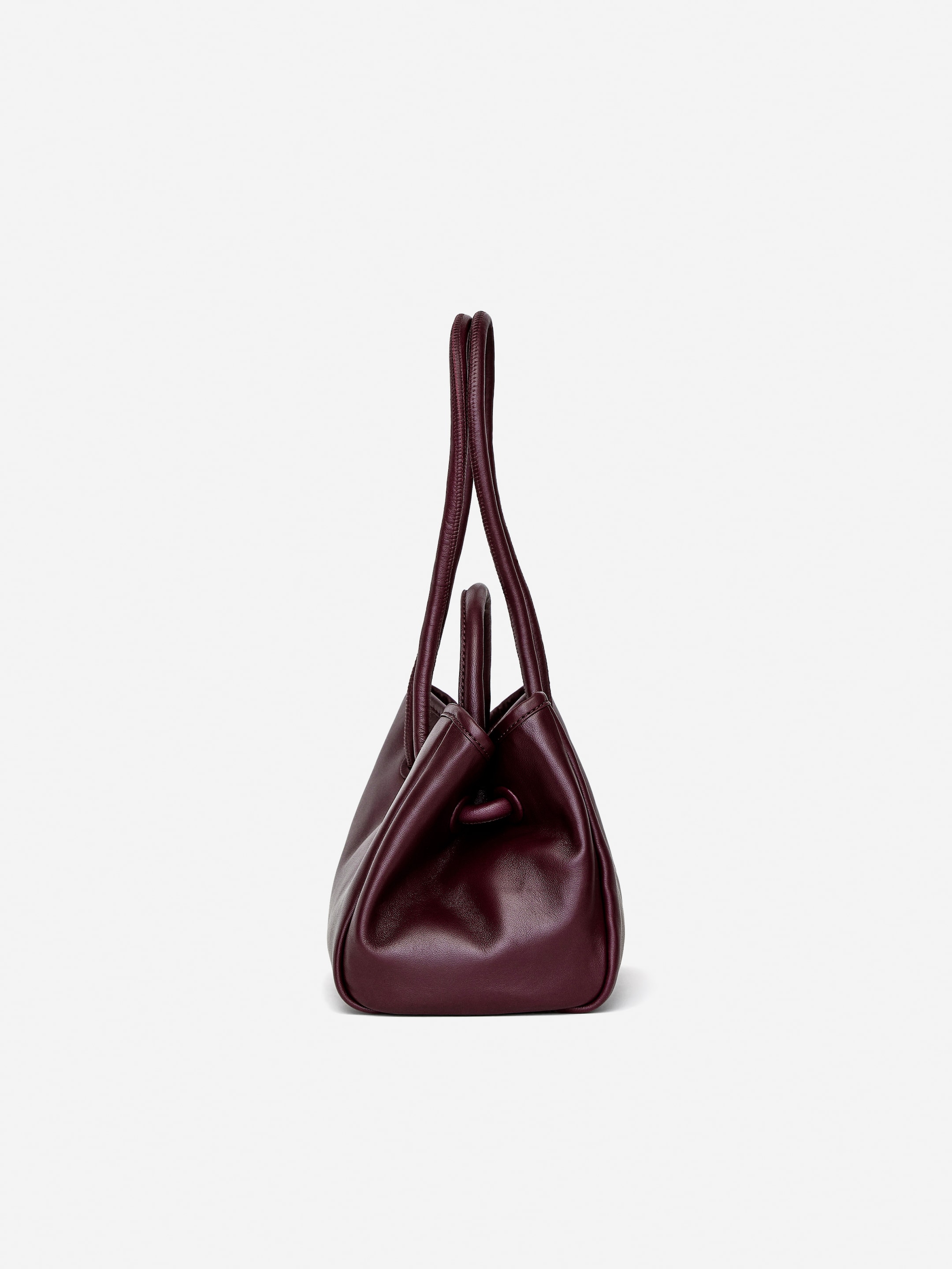 Visa större bild: En vinröd läder crossbody-väska står vinklad åt vänster, med runda topphandtag som är integrerade i dess mjuka kropp genom rynkade öljetter.