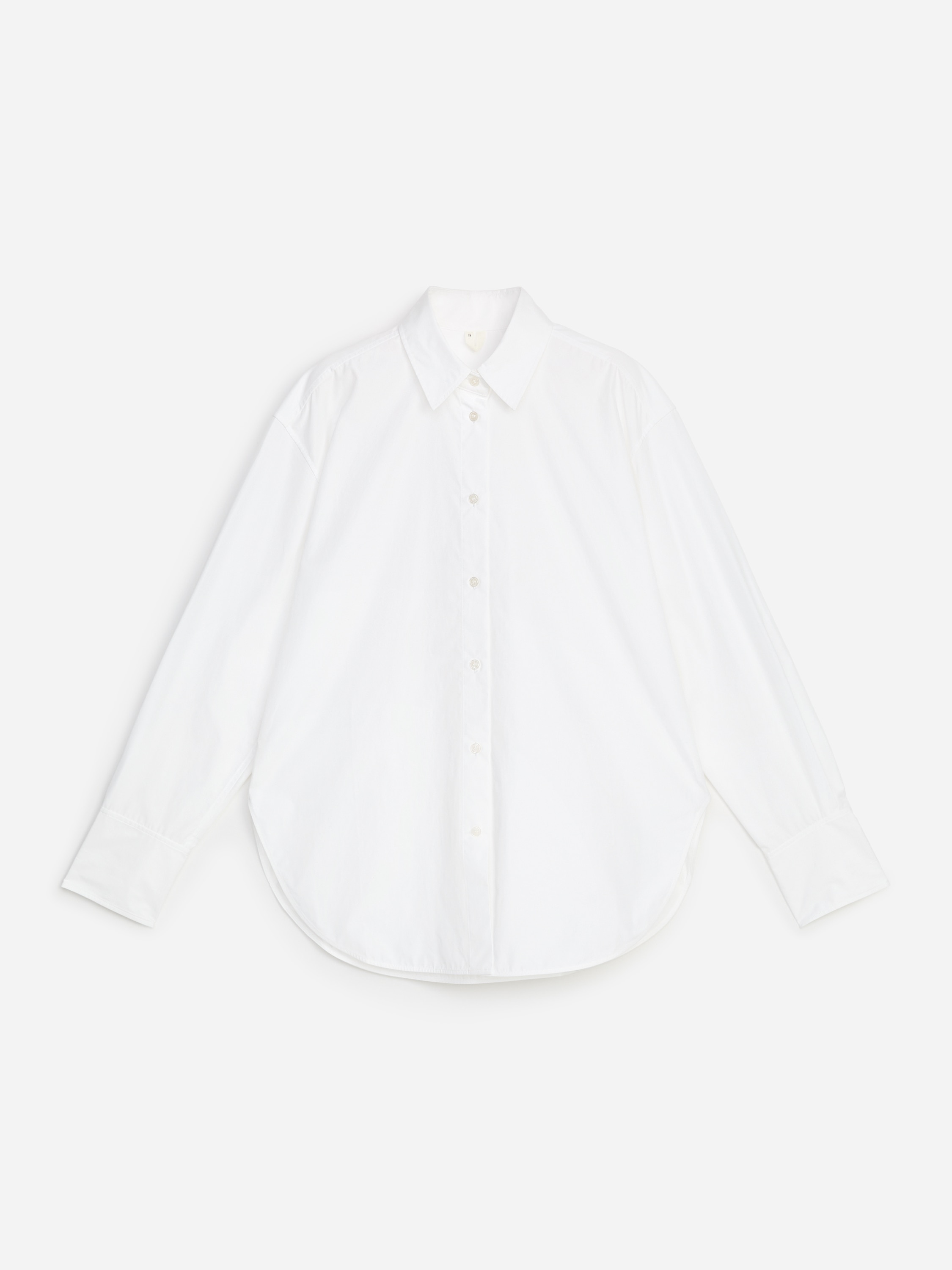 A white, long-sleeved button-up shirt in crisp cotton poplin is worn by the model, featuring a relaxed silhouette, a pointed collar, and a curved hem. The shirt is styled with the top buttons undone and the sleeves slightly pushed up, paired with black trousers.