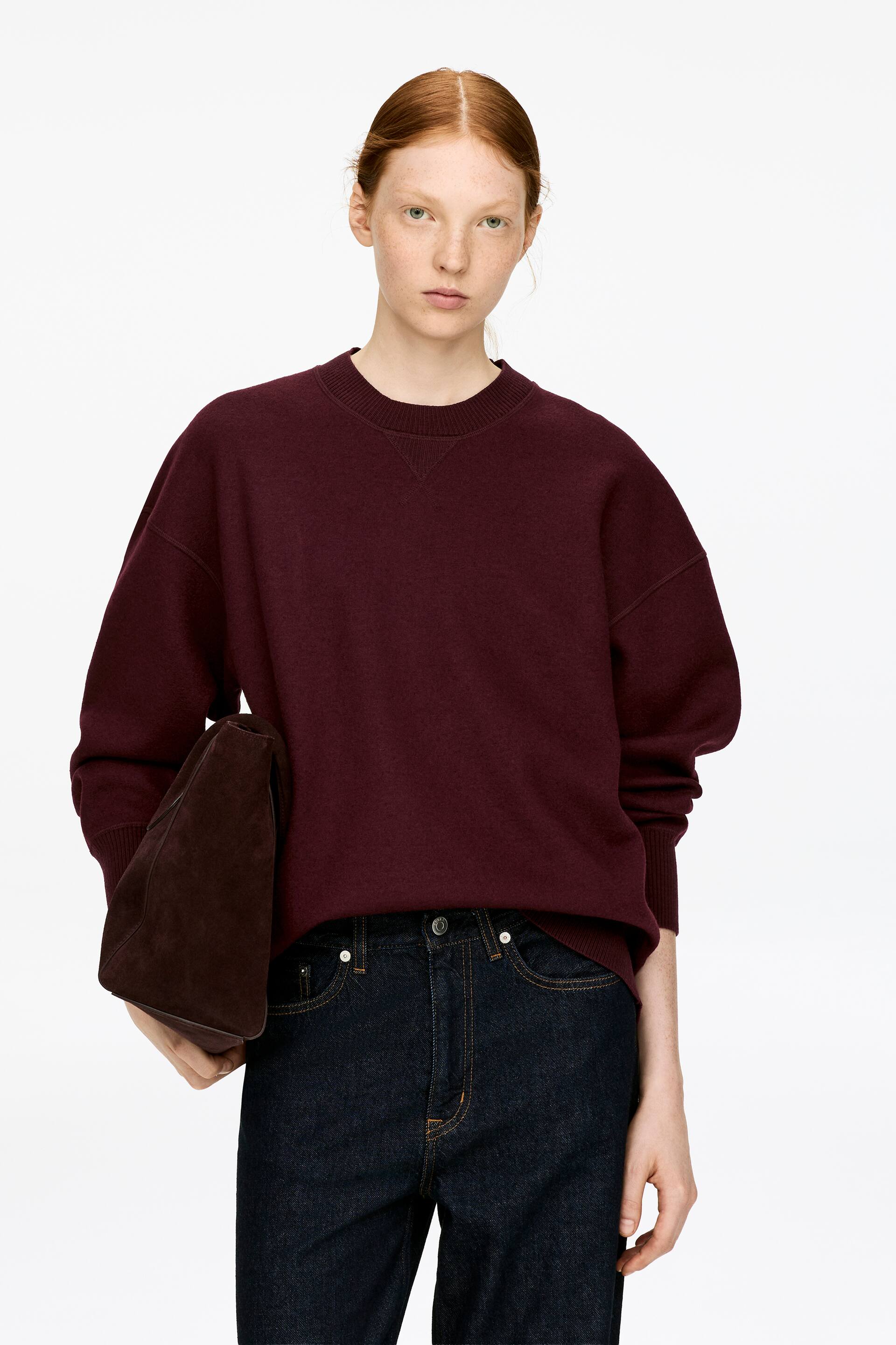 Vis større bilde: En vinrød genser med rundt hals, med en oversized passform med droppede skuldre og ribbestrikkede kanter i halsen, ved mansjettene og nederst, bæres av modellen. Modellen holder også en mørkebrun veske i en hånd.