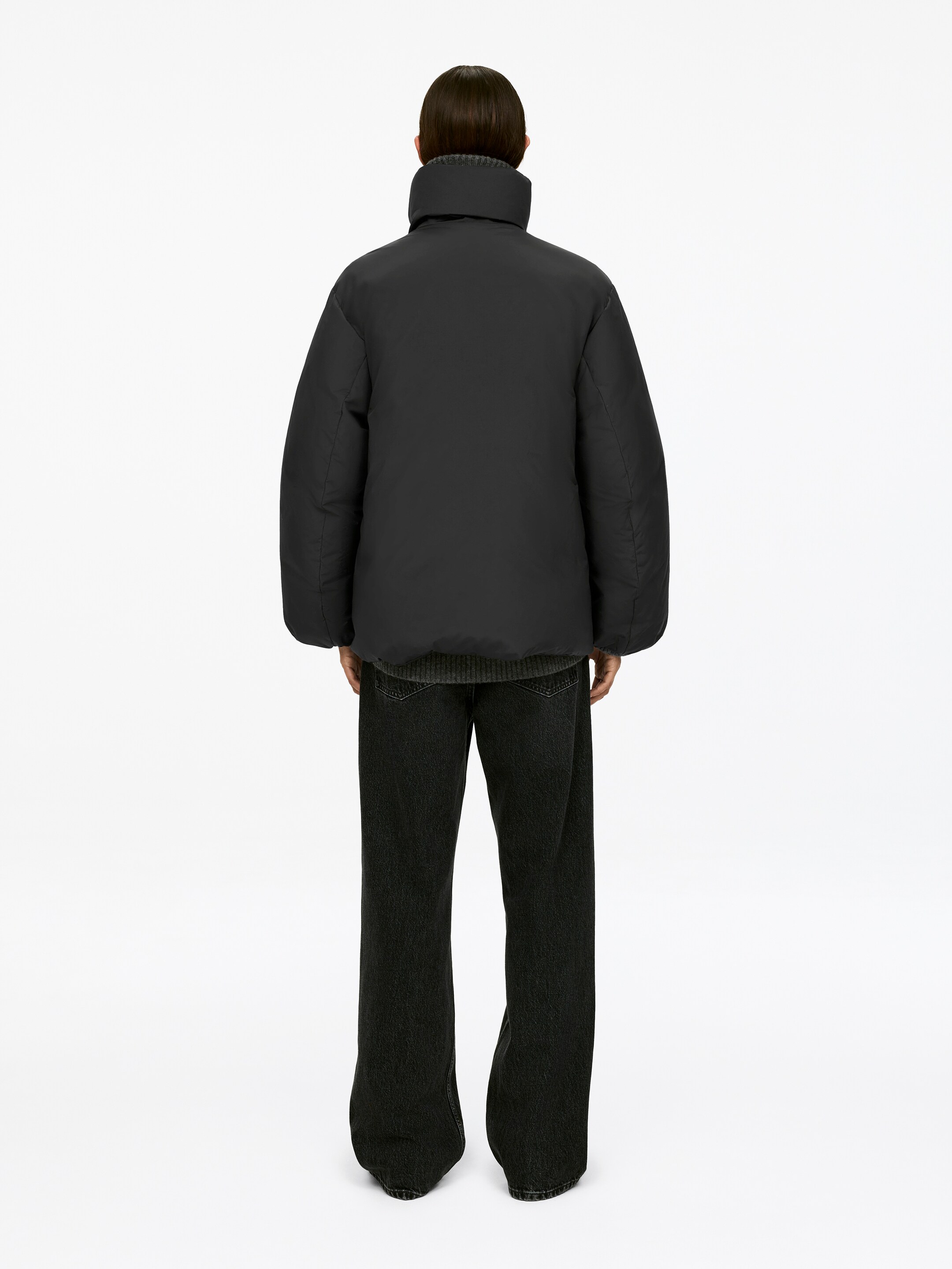 Größeres Bild ansehen: Eine schwarze Daunenjacke, die einen Stehkragen aufweist, wird von dem Modell getragen, das von hinten zu sehen ist. Ein grauer gerippter Pullover ist darunter sichtbar, und dunkle Hosen mit weitem Bein werden ebenfalls getragen.