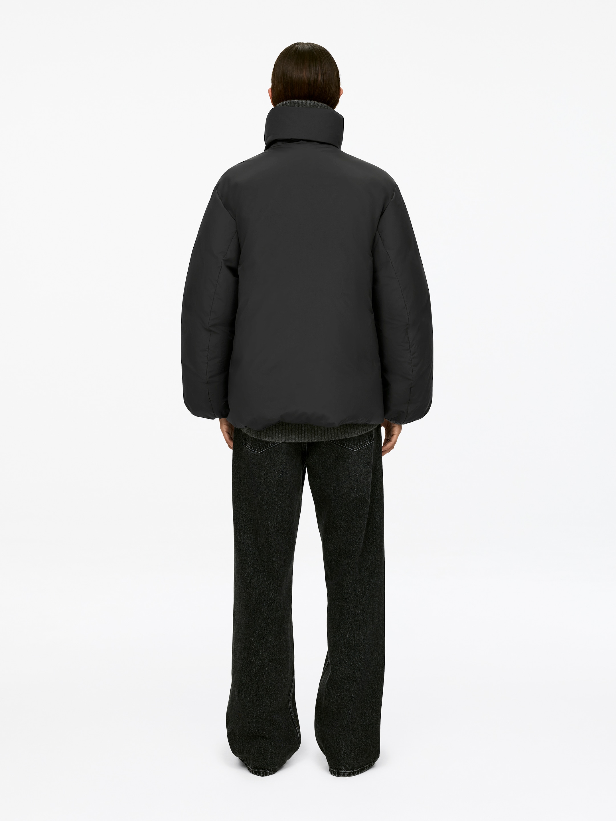 Agrandir l'image: Une doudoune noire, avec un col montant, est portée par le mannequin, vue de dos. Un pull gris à côtes est visible en dessous, et un pantalon large et sombre est également porté.