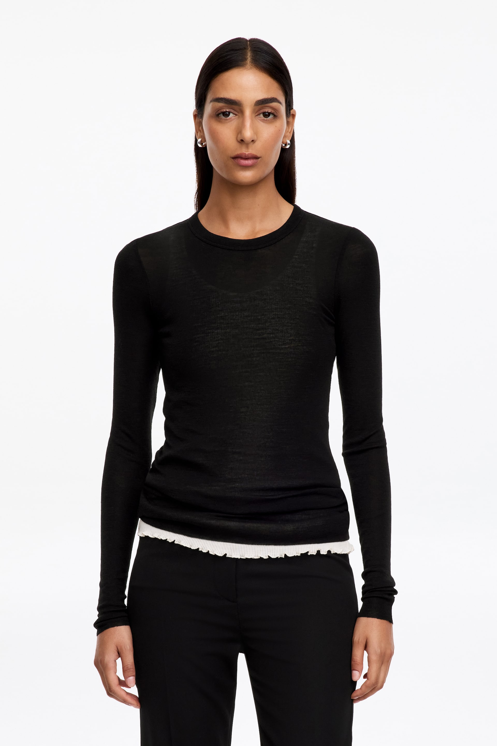 Agrandir l'image: Top en mérinos à manches longues. Un top noir en mérinos à maille fine, à manches longues et à col rond, est porté par le mannequin. Son tissu présente une texture légèrement transparente, et un ourlet blanc à volants est visiblement superposé sous son bord inférieur.