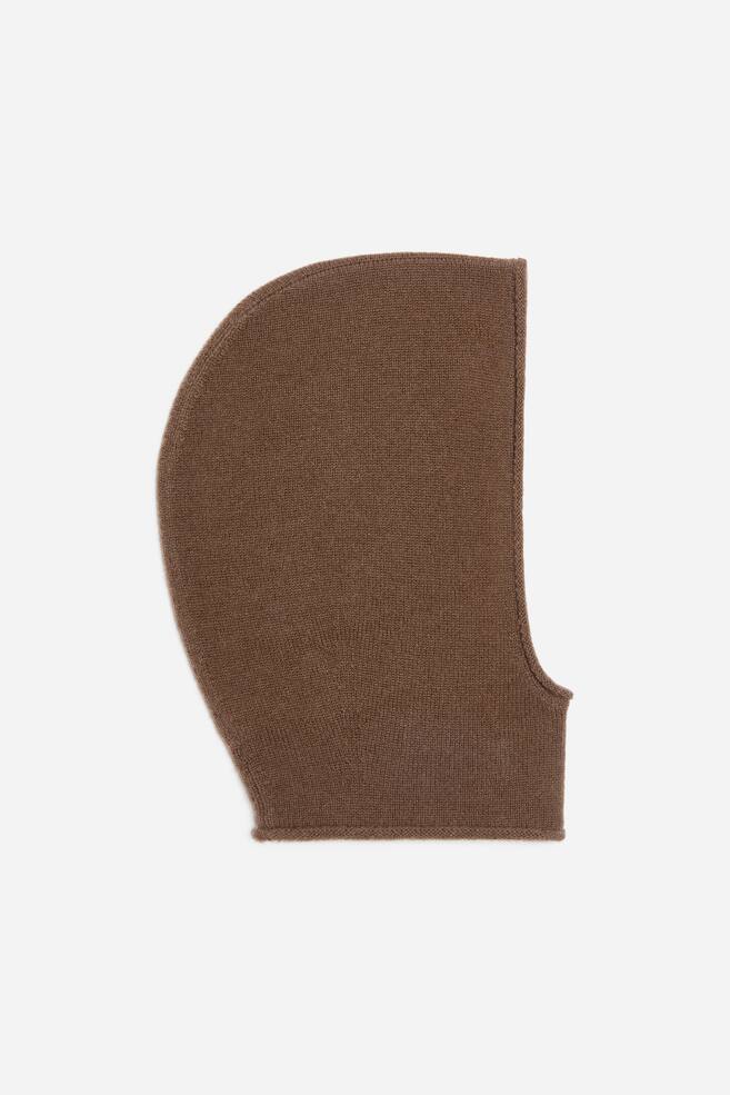 A brown knitted balaclava is lying flat in side profile, facing right, showcasing its curved hood silhouette and fine-knit texture. The balaclava features a vertical face opening and clean-finished edges around the face and neck. - 1