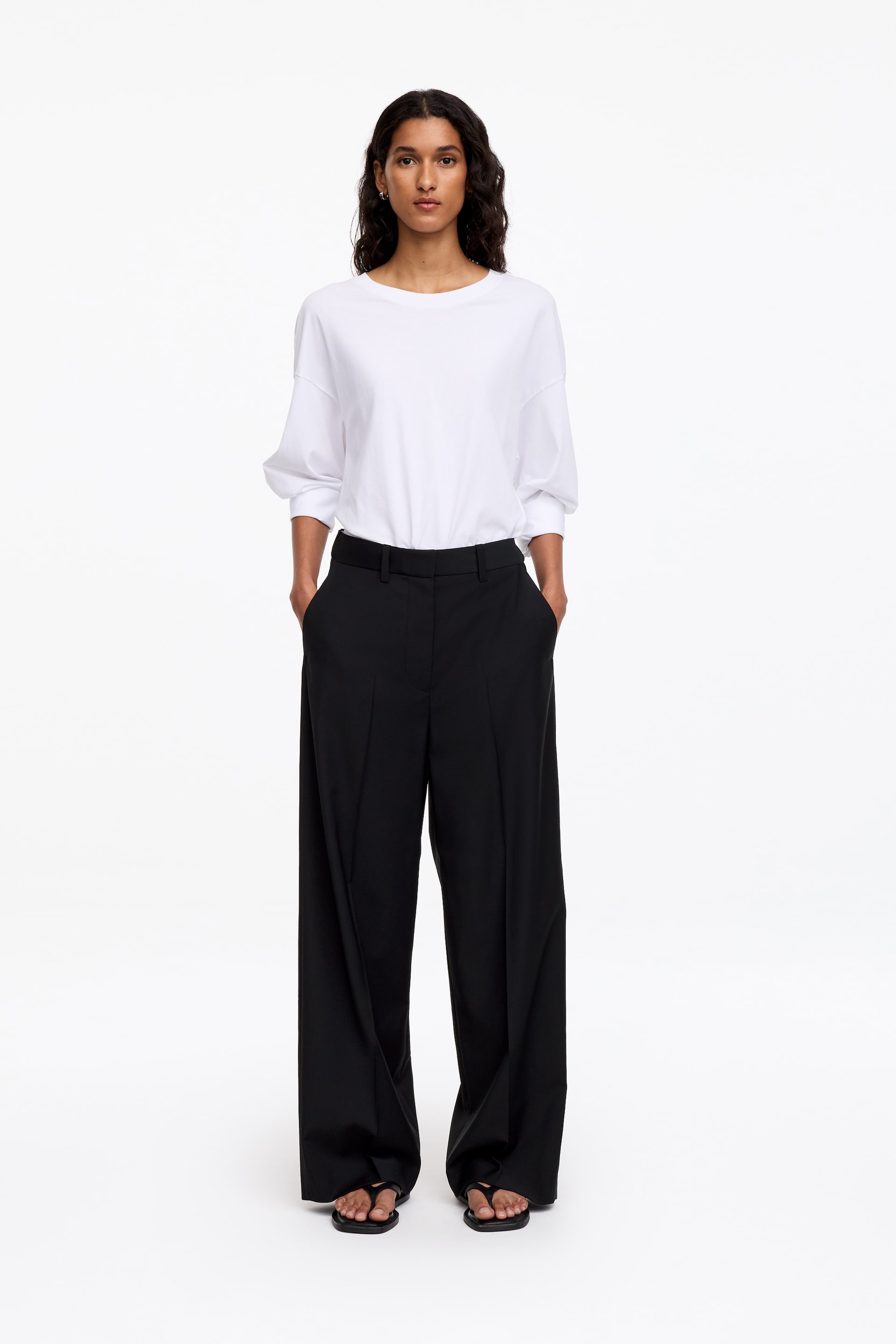 Visualizza l'immagine ingrandita: Questi pantaloni neri sartoriali, caratterizzati da una silhouette ampia e rilassata e da pieghe frontali marcate, sono indossati dalla modella, in piedi con le mani appoggiate nelle tasche laterali. I pantaloni sono abbinati a un top bianco a maniche lunghe infilato dentro e a sandali neri bassi.