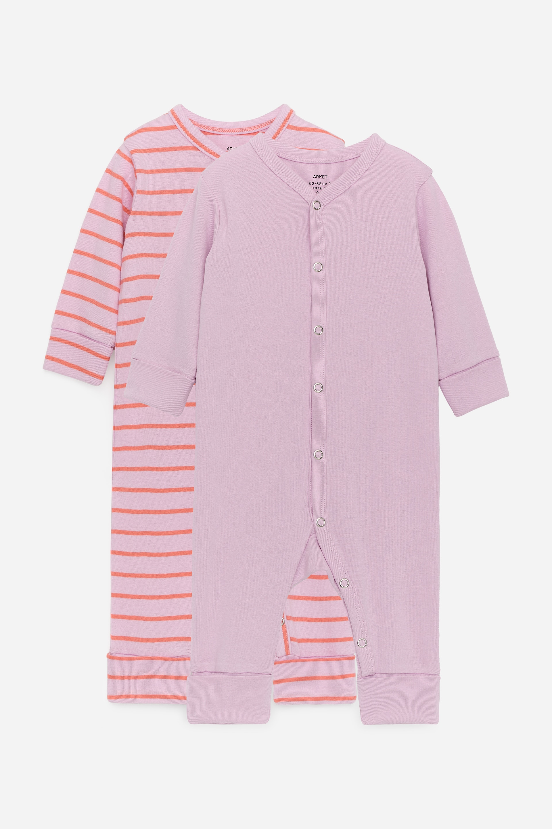 View larger image: A set of two pyjama overalls is on display. One overall is a solid light purple, while the other is light pink with horizontal orange stripes, both featuring long sleeves and legs with foldable cuffs. The purple overall showcases snap fastenings running down its front from top to toe.