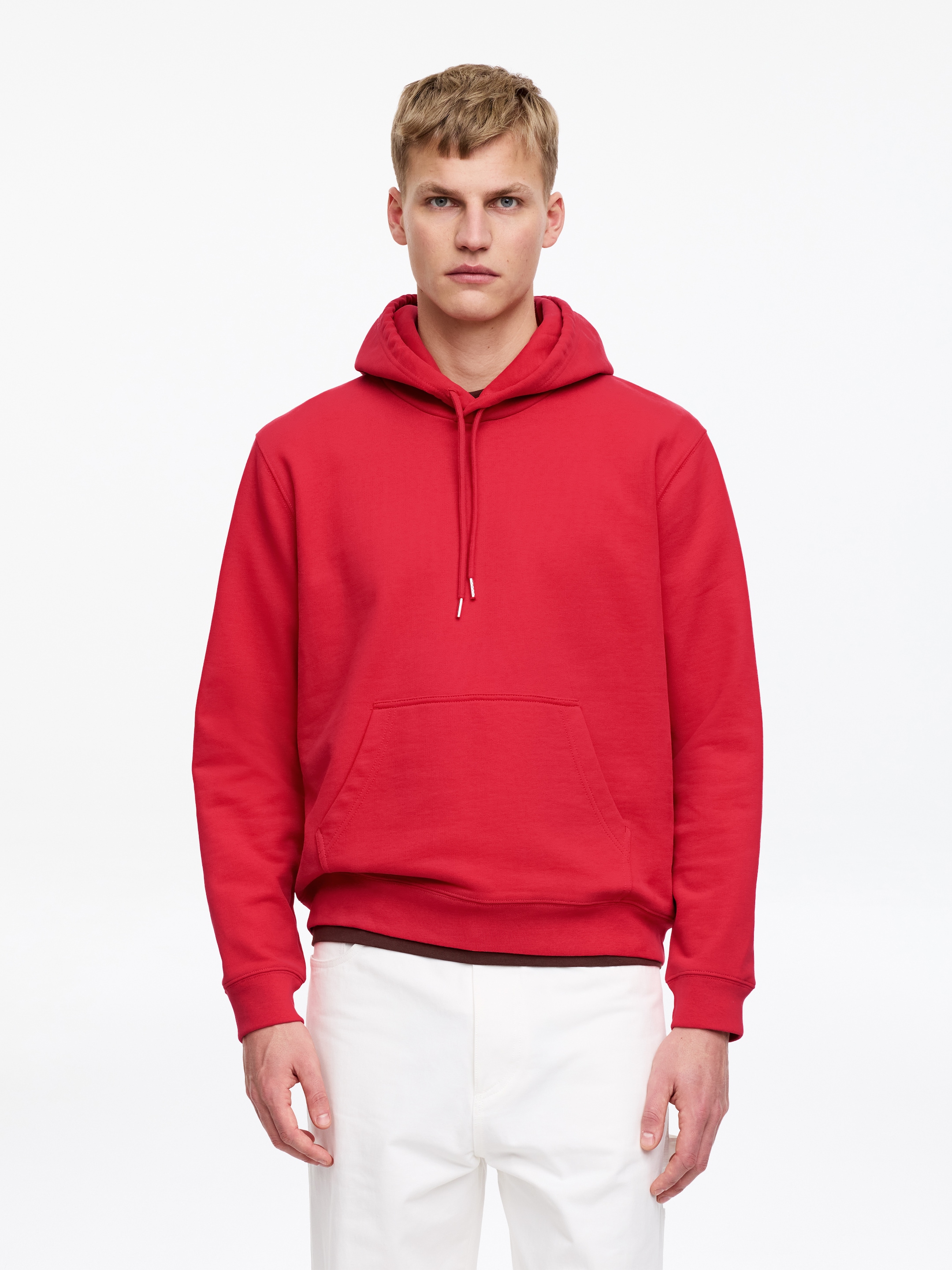 Ver imagem maior: Uma sweatshirt vermelha com capuz, com cordões, bolso canguru frontal e punhos e bainha canelados, é usada pelo modelo num corte regular, sobre uma t-shirt escura e com calças brancas.