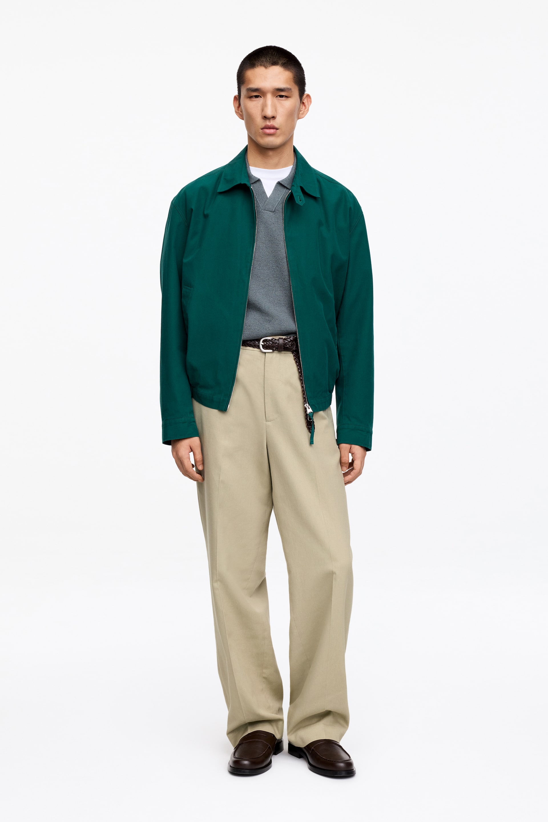 Agrandir l'image: Une veste vert foncé à col avec une fermeture éclair sur le devant est portée par-dessus un pull gris à col en V et un t-shirt blanc. Un pantalon kaki à jambes larges est porté, ajusté avec une ceinture tressée marron, accompagné de mocassins marron foncé.