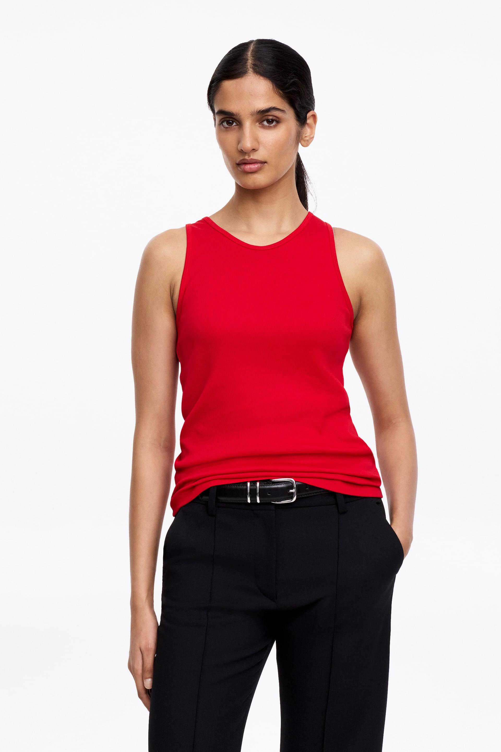 Vis større bilde: En rød ribbestrikket tanktopp med rund hals bæres av modellen. Den er stylet ned i svarte bukser, som er sikret med et svart belte.
