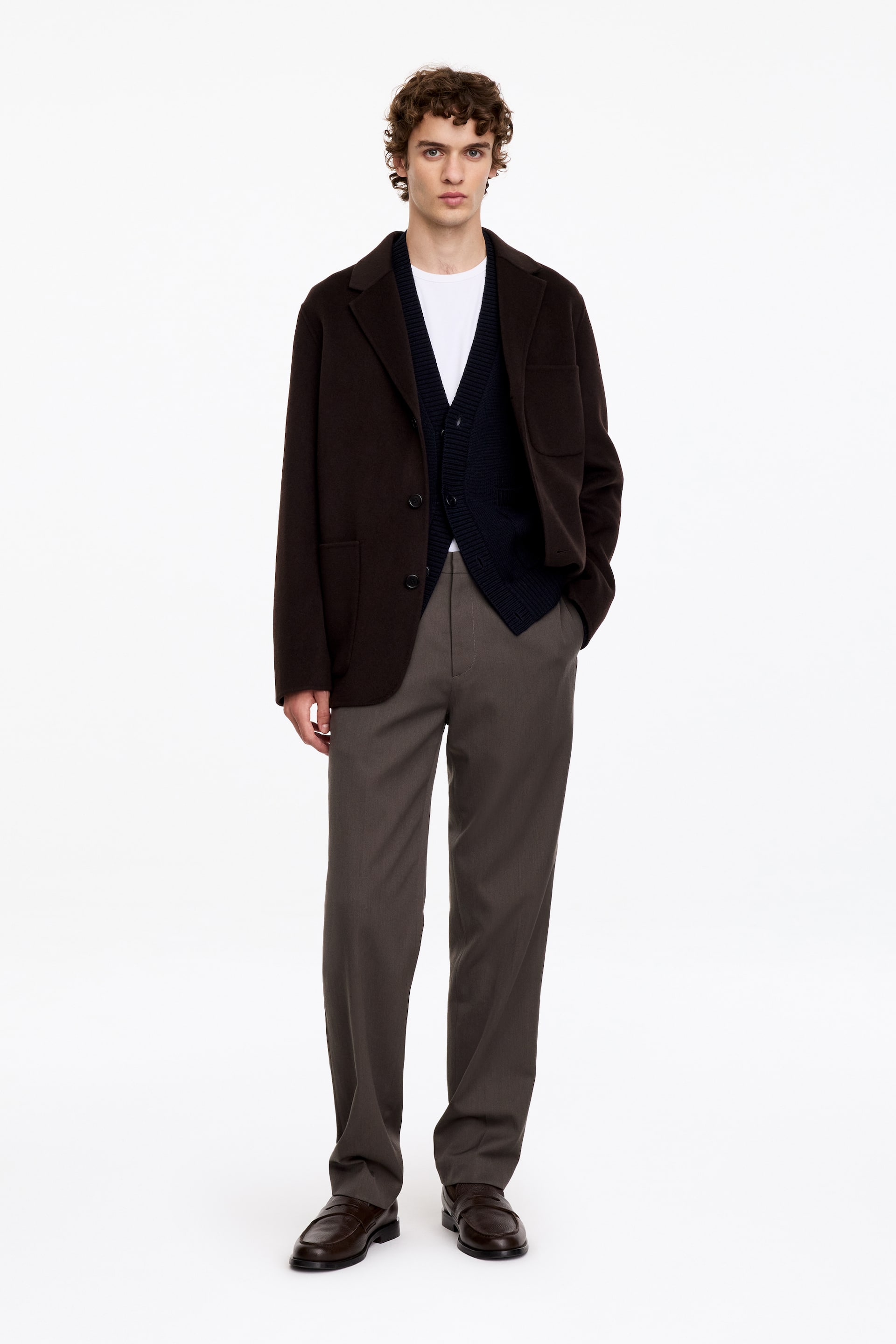 Visualizza immagine più grande: I pantaloni in misto lana e cotone in una tonalità grigio-marrone scuro sono indossati dalla modella e presentano una silhouette a gamba dritta e una fascia in vita pulita. I pantaloni sono abbinati a una t-shirt bianca, un cardigan in maglia blu navy, un blazer marrone scuro e mocassini in pelle marrone scuro.