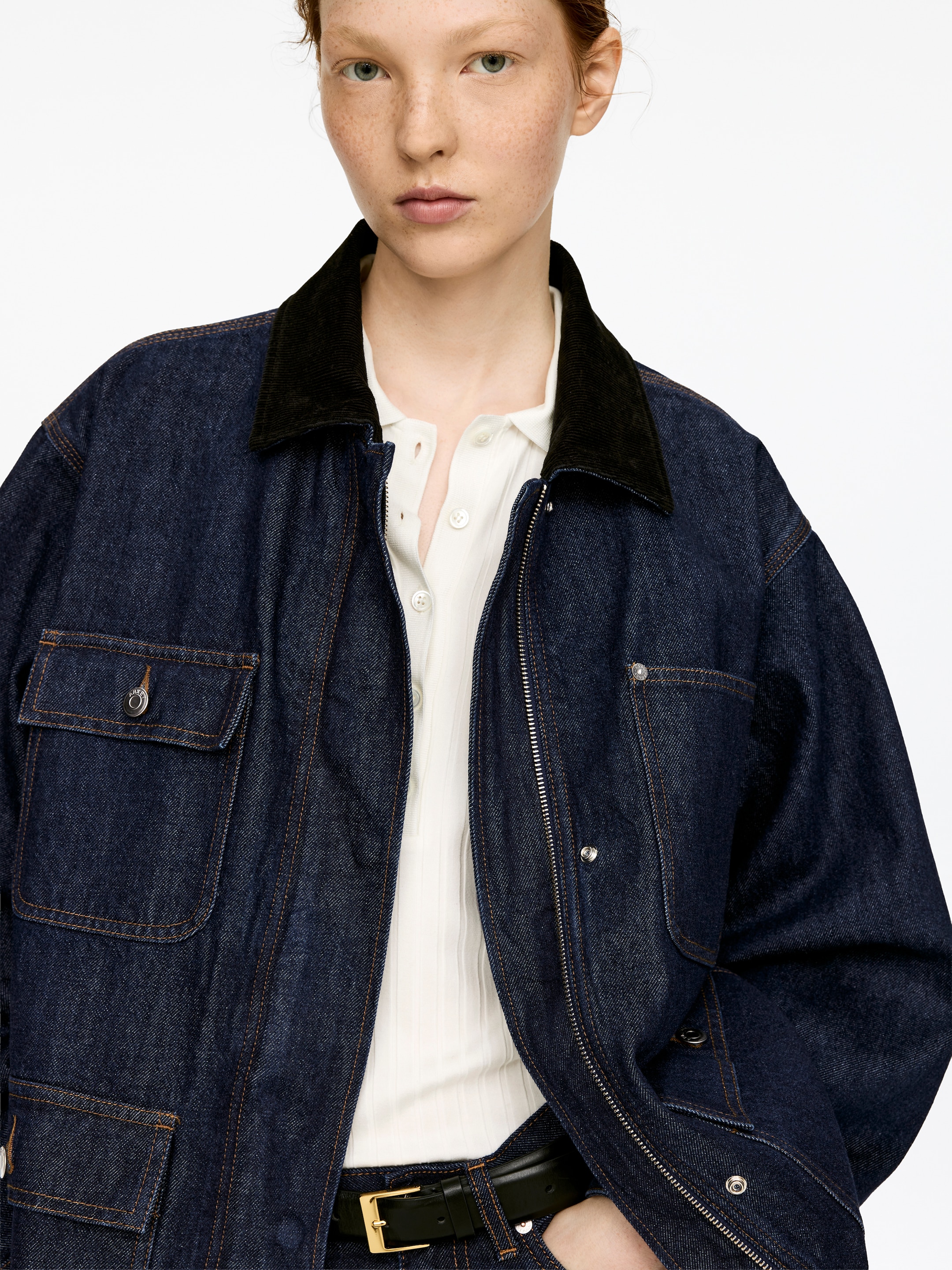 Visualizza immagine più grande: Una giacca di jeans blu scuro oversize con un colletto in velluto a coste nero è indossata dal modello. Presenta una chiusura metallica visibile con zip e diverse tasche anteriori applicate, alcune con patte abbottonate, stratificate sopra una camicia a coste bianca e una cintura nera.