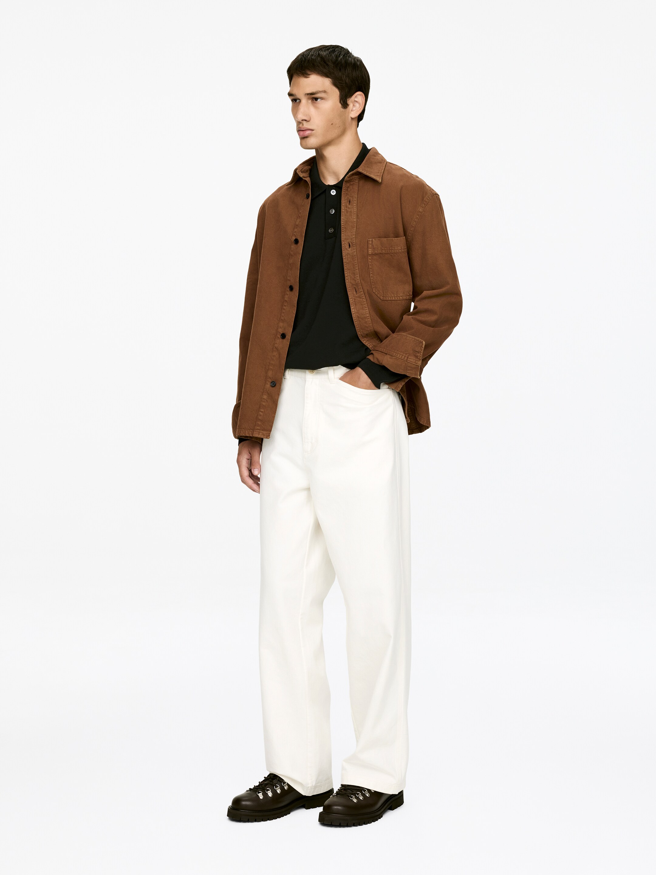 Visualizza immagine più grande: Una camicia marrone a maniche lunghe con colletto è portata aperta sopra una polo nera a maniche lunghe. Un paio di pantaloni larghi color avorio e scarpe scure stringate, con occhielli a D-ring e suole carrarmato, completano l'insieme, che viene presentato da una vista frontale a figura intera leggermente angolata.
