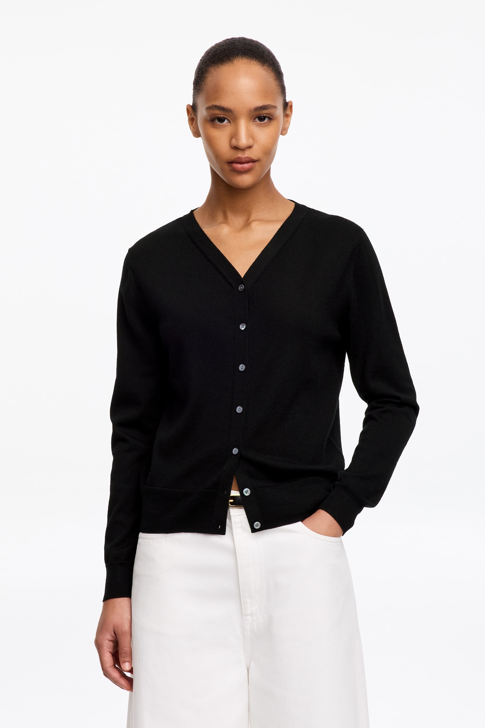 Vezi imaginea mai mare: Un cardigan negru din lână Merino evidențiază un decolteu în V și nasturi frontali de culoare deschisă. Este purtat de model peste pantaloni albi, iar o parte dintre nasturi sunt prinși.