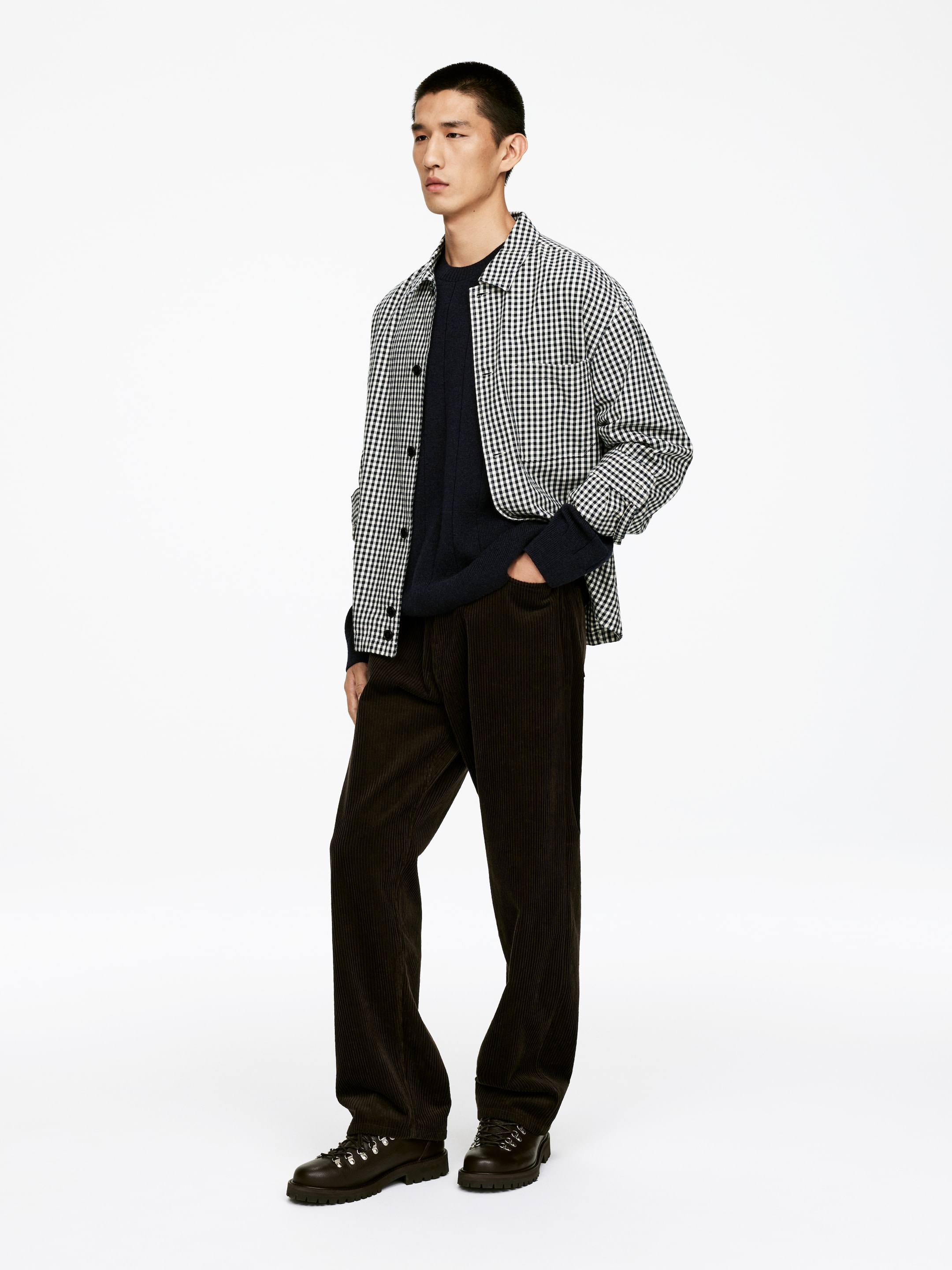 View larger image: A black and white checkered long-sleeved shirt is being worn open over a dark navy ribbed crewneck sweater, presented from a three-quarter angle. Dark brown wide-leg corduroy pants are being worn, along with dark hiking boots featuring metal eyelets and thick lug soles.