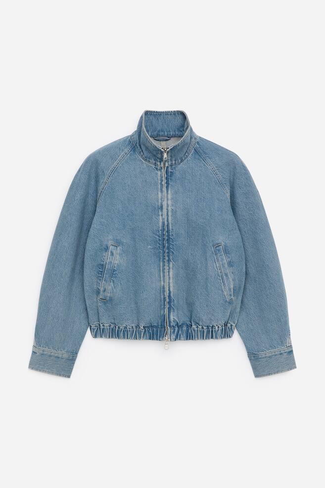Eine kurze Jeansjacke in hellblauer Baumwolle präsentiert ihre FRONT, definiert durch einen elastischen Bund und gedrehte Ärmel. Sie verfügt über einen Trichterkragen, einen zentralen Reißverschluss und zwei schräge Fronttaschen. - 1