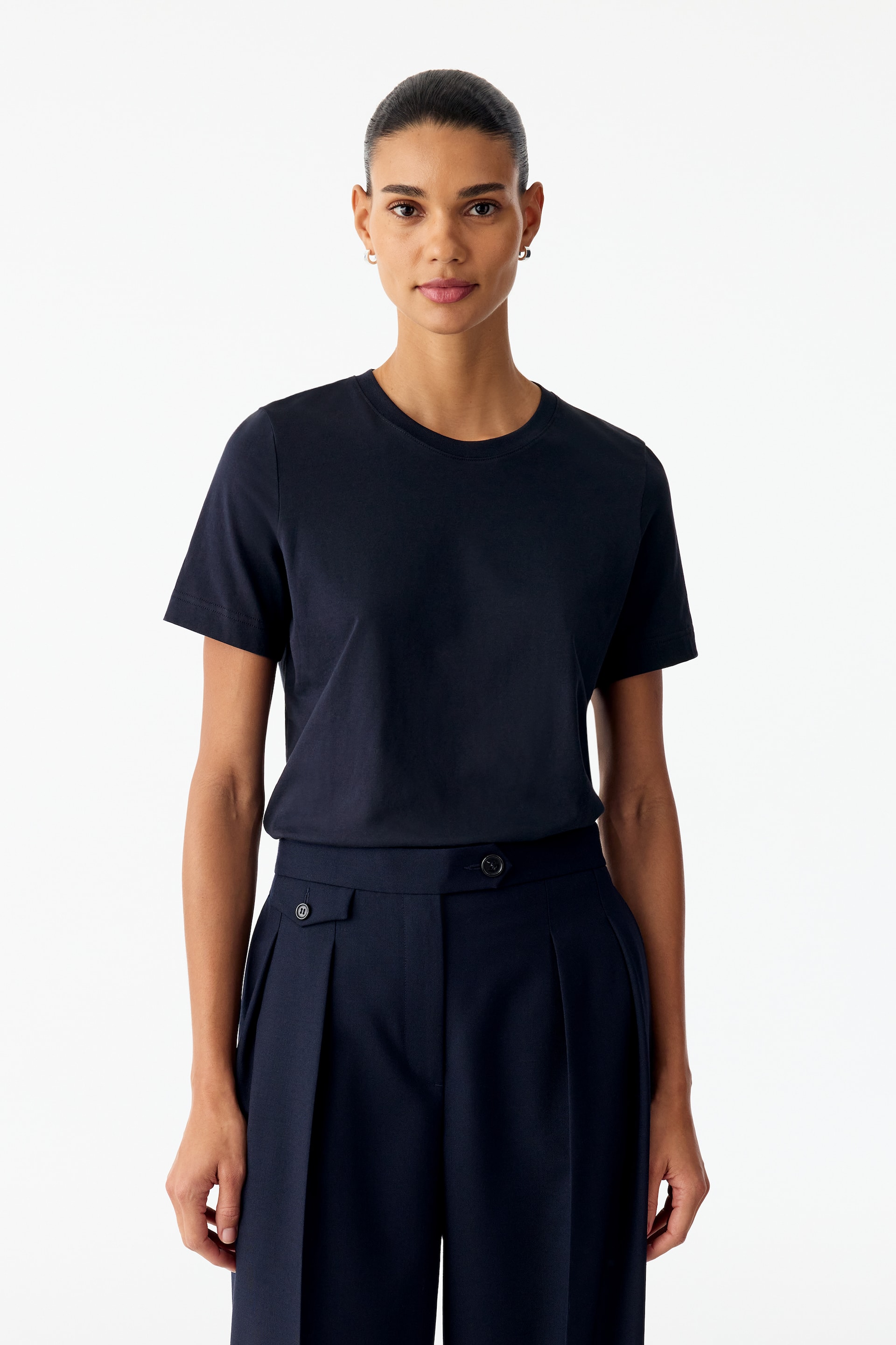 Visualizza l'immagine ingrandita: Una T-shirt girocollo blu navy scuro con maniche corte è indossata dalla modella, infilata dentro pantaloni pieghettati a vita alta dello stesso colore. La T-shirt presenta una vestibilità regular classica e un colletto a costine.