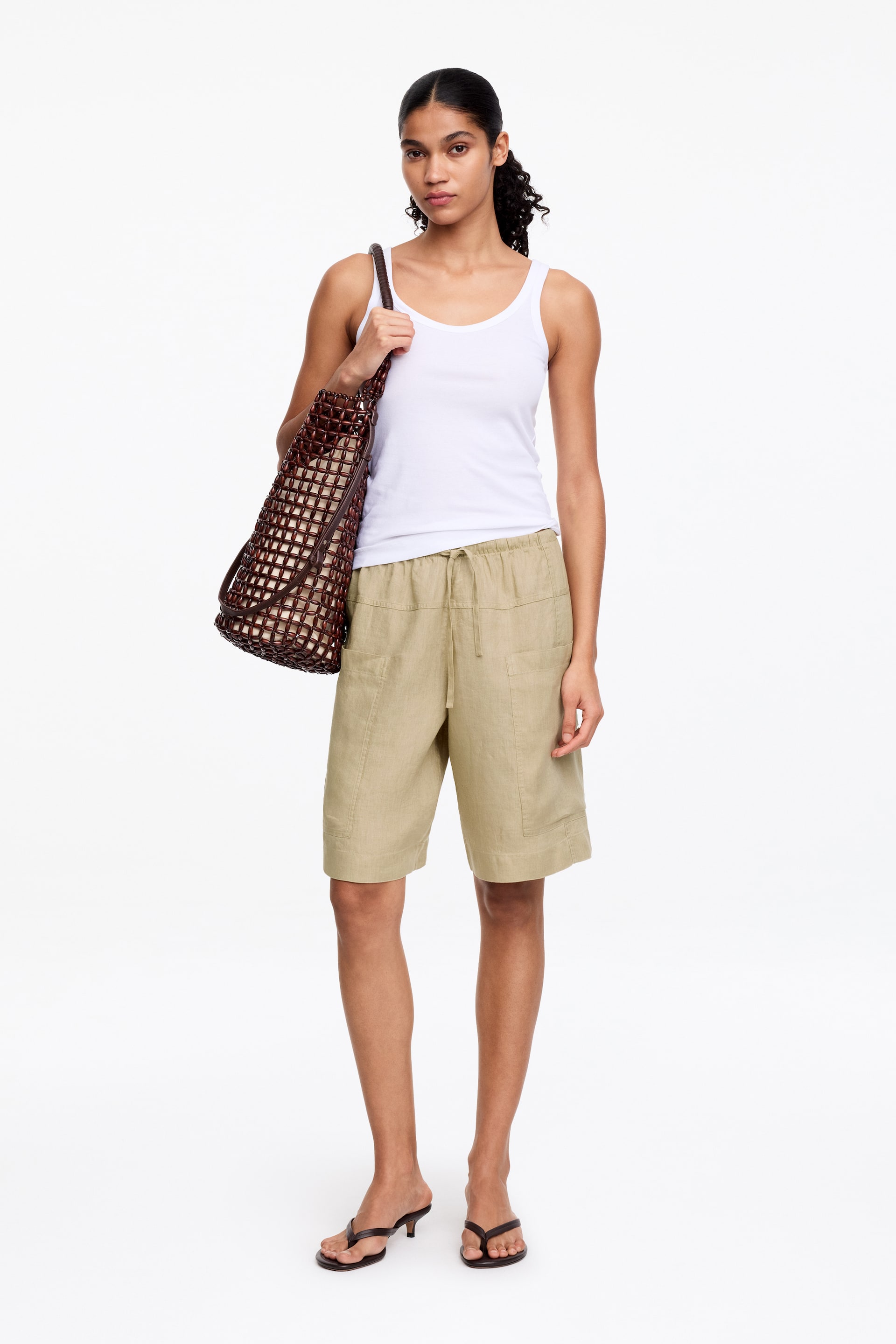 Se større billede: Outfittet består af en hvid tanktop med rund halsudskæring, khaki shorts med snøre i taljen og påsyede lommer samt brune kitten heels i flip-flop stil. En brun flettet tote taske med mønstret yderside bæres på højre skulder.