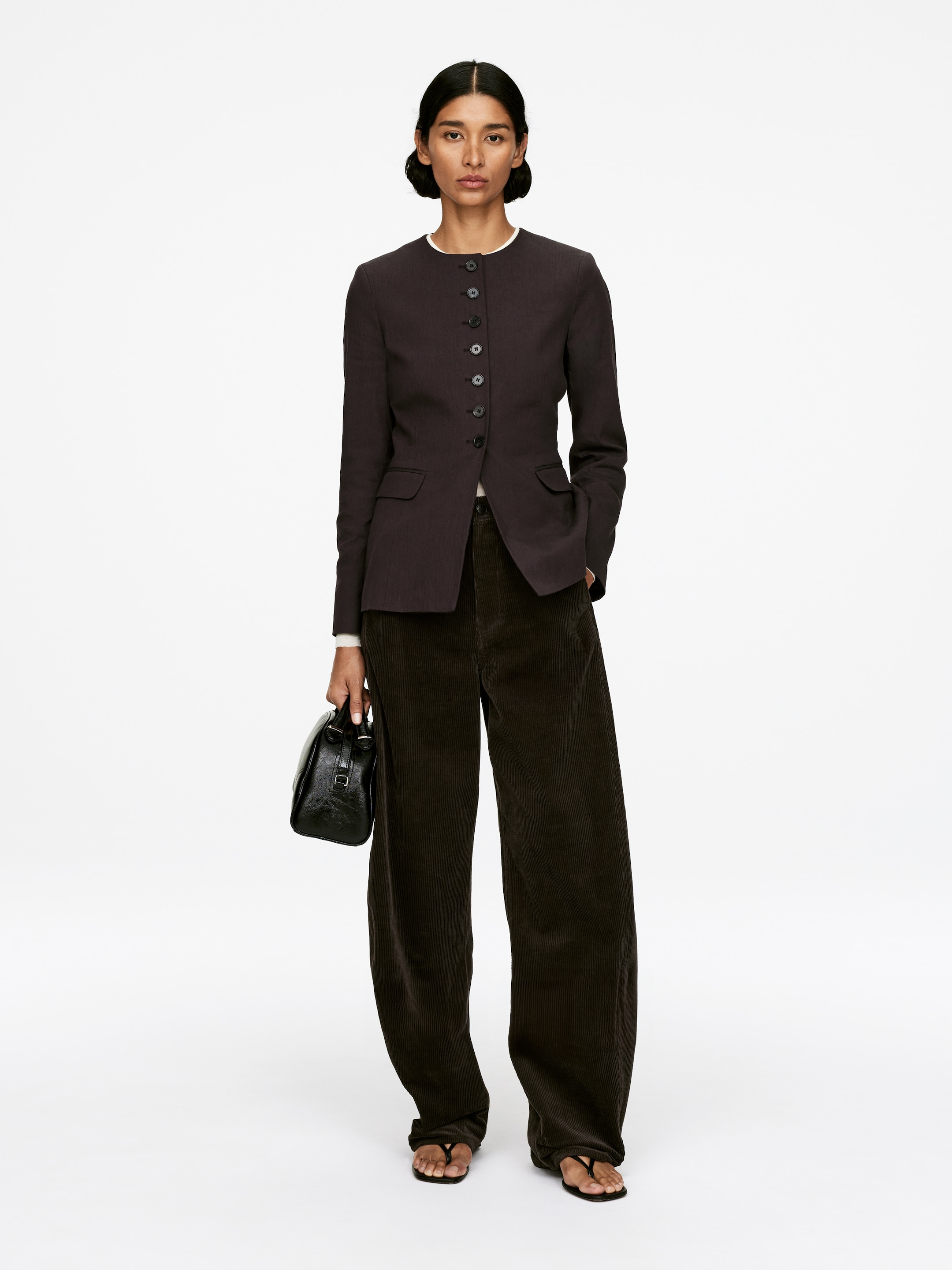 View larger image: A front-facing full outfit is being presented, featuring a dark charcoal collarless jacket with a subtle pinstripe pattern, a full-length button placket, and two flap pockets, worn over a white top. Dark brown wide-leg corduroy trousers are being styled with the jacket, pooling at the ankles, along with black thong sandals, and a small black structured top-handle handbag is being held, completing the ensemble.