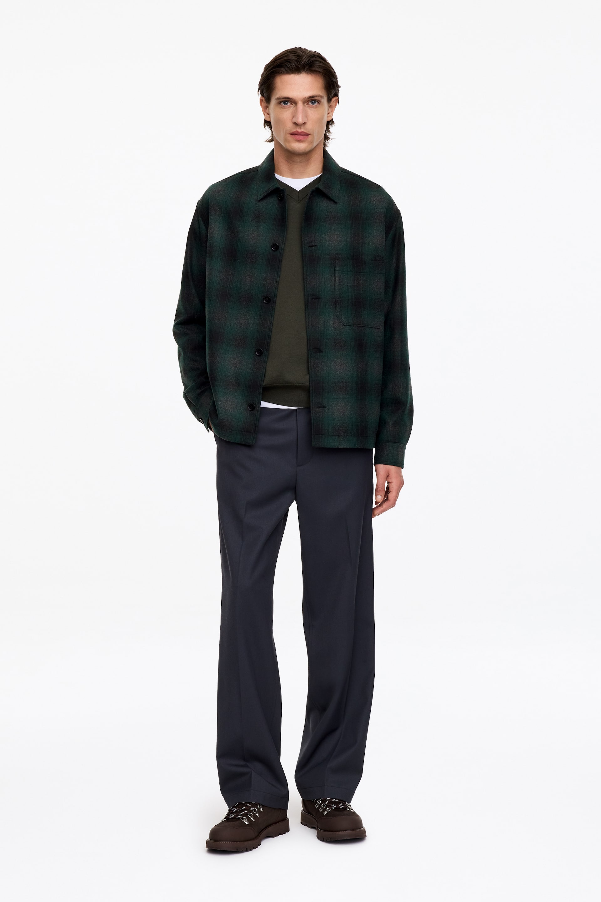 Visualizza l'immagine ingrandita: La modella indossa aperta una overshirt a quadri verde e nera, con vestibilità rilassata, colletto classico e una tasca sul petto. L’overshirt è indossata sopra un pullover verde scuro con scollo a V e una T-shirt bianca, abbinata a pantaloni grigio scuro.