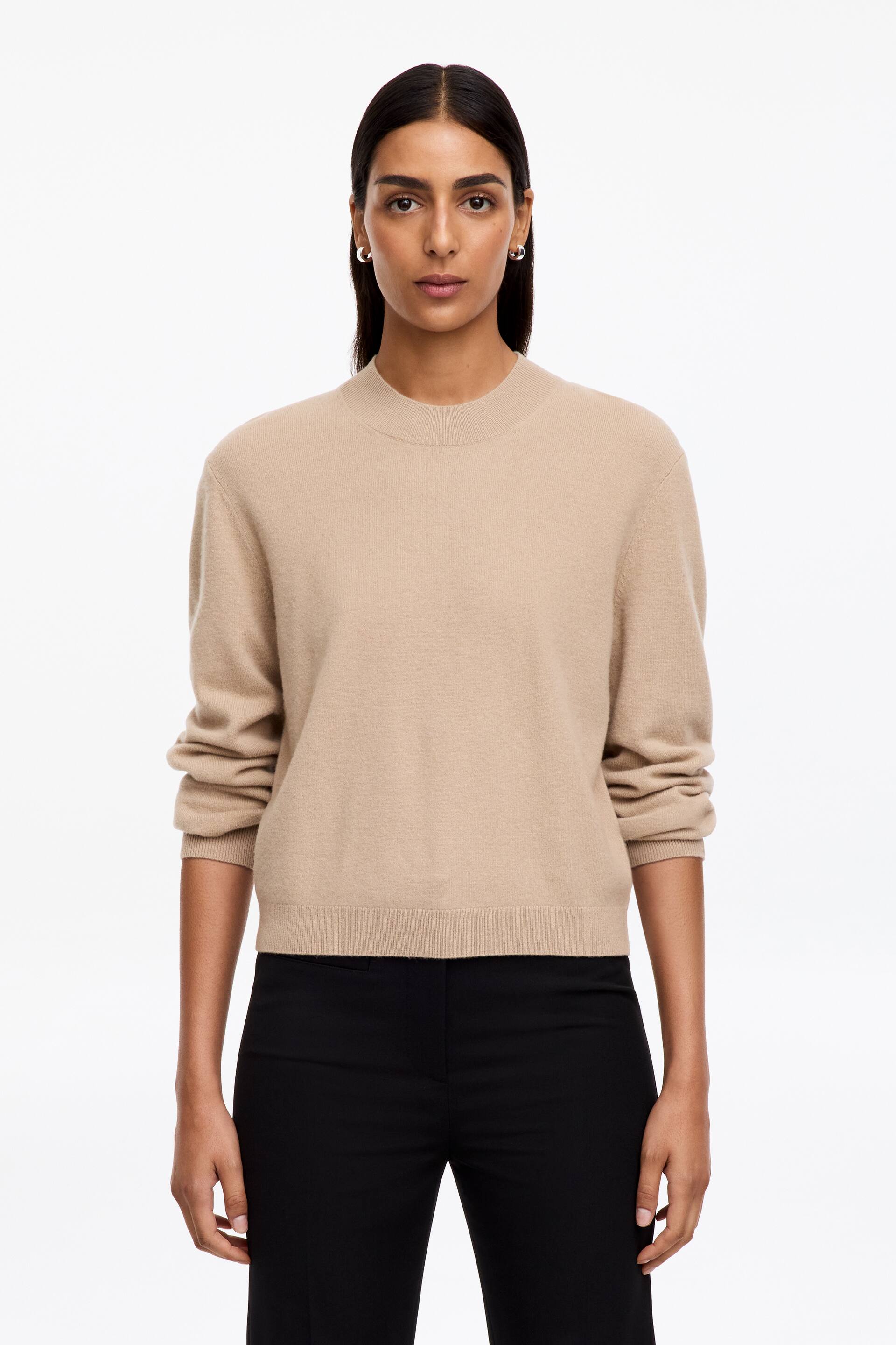 Agrandir l'image: Un pull en maille beige à col rond, avec poignets et ourlet côtelés, est porté par le mannequin. Le pull est ajusté près du torse du mannequin et est associé à un pantalon noir.
