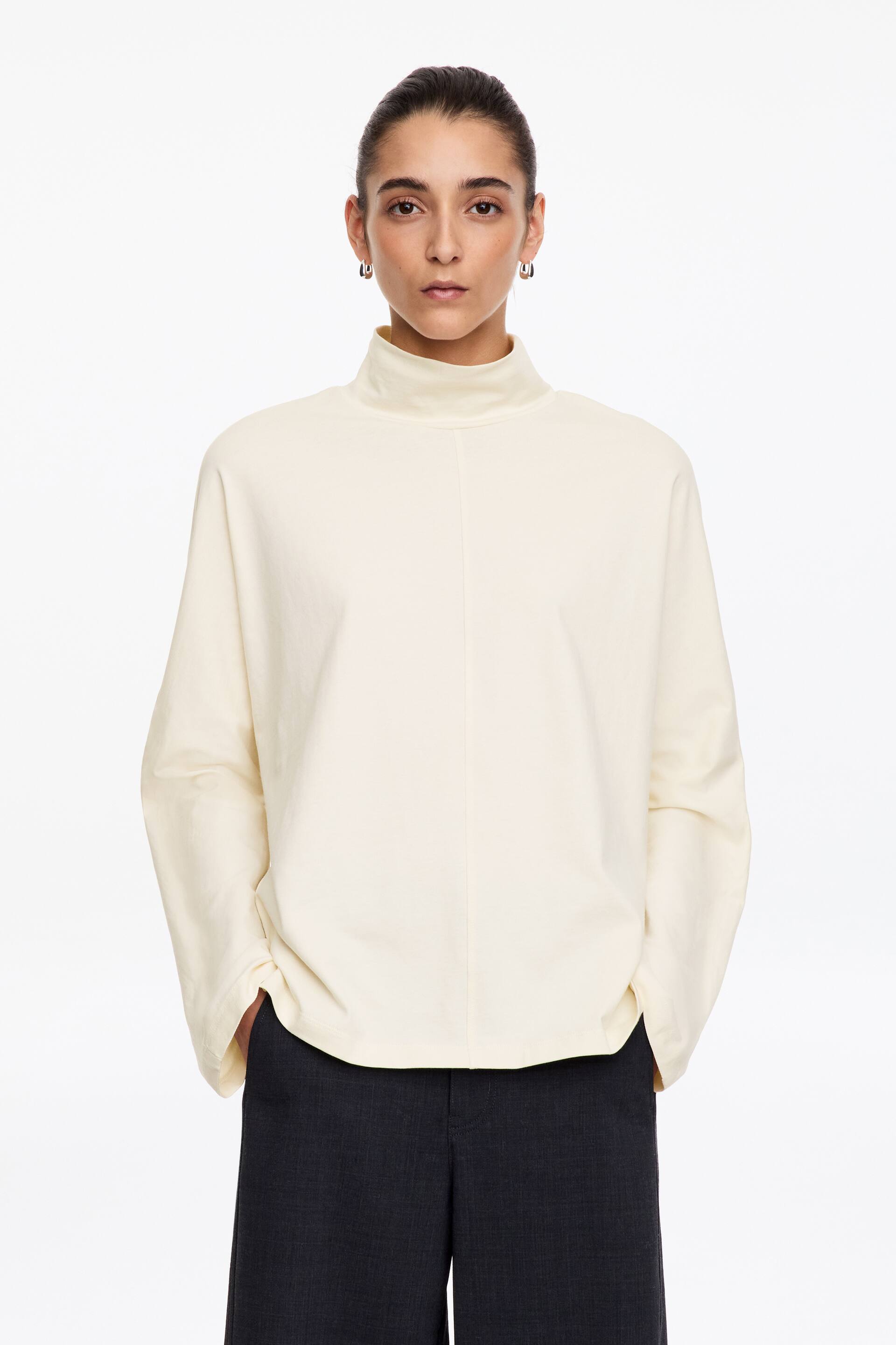 View larger image: An off-white, relaxed-fit top featuring a high roll neck, long batwing sleeves, and a vertical centre-front seam is being worn by the model. The model is standing with her hands tucked into the pockets of dark grey trousers.
